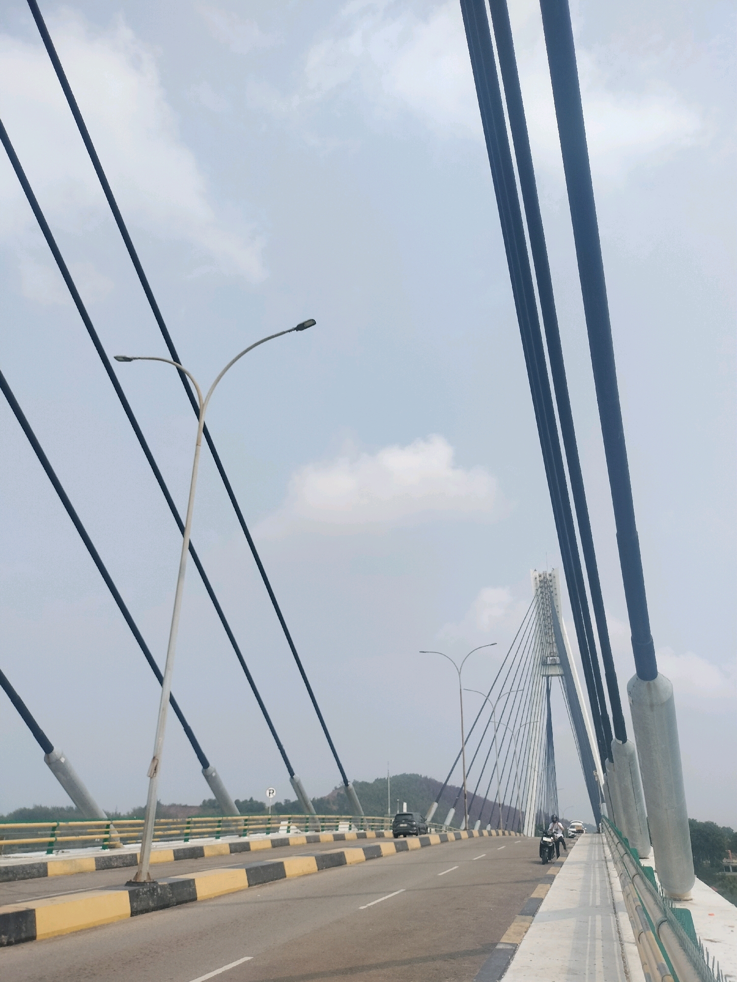 The image depicts a view of the cable-stayed bridge, likely the Batam-Bintan Bridge, in Batam, Indonesia. The perspective is from the roadway of the bridge, looking towards the main tower. Several dark cables extend diagonally from the unseen main tower towards the bridge deck. A white lamppost with two lamps is visible on the left side of the road, positioned between two of the main cables. The road has clear lane markings and a yellow and black striped barrier along the side. In the distance, a few vehicles, including cars and a motorcycle with two riders, are seen on the bridge. The sky is partly cloudy and the background shows a hilly landscape with vegetation. The overall scene is characterized by the imposing structure of the bridge against a natural backdrop.