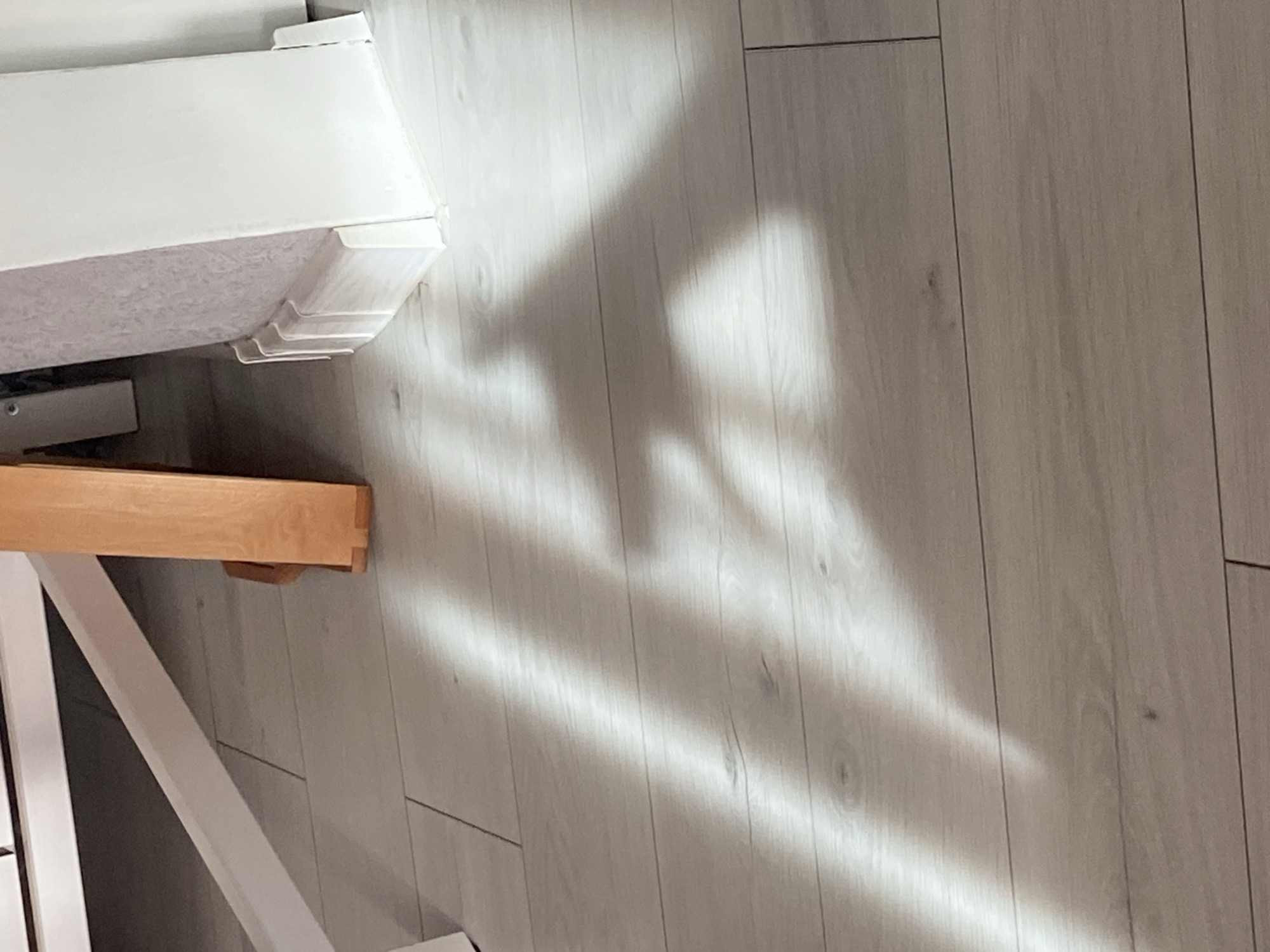 The image depicts a floor with light and shadow patterns. The floor is made of light-colored wooden planks, arranged horizontally. A prominent shadow, cast by an unseen object, creates a shape resembling a heart on the floor. The shadow is partially illuminated by streaks of light, suggesting sunlight entering from an angle. In the background, parts of furniture and a white baseboard are visible. The overall scene is indoors, with a focus on the floor and the interplay of light and shadow. There is no discernible information about the city of Brest, Belarus, within the image itself.