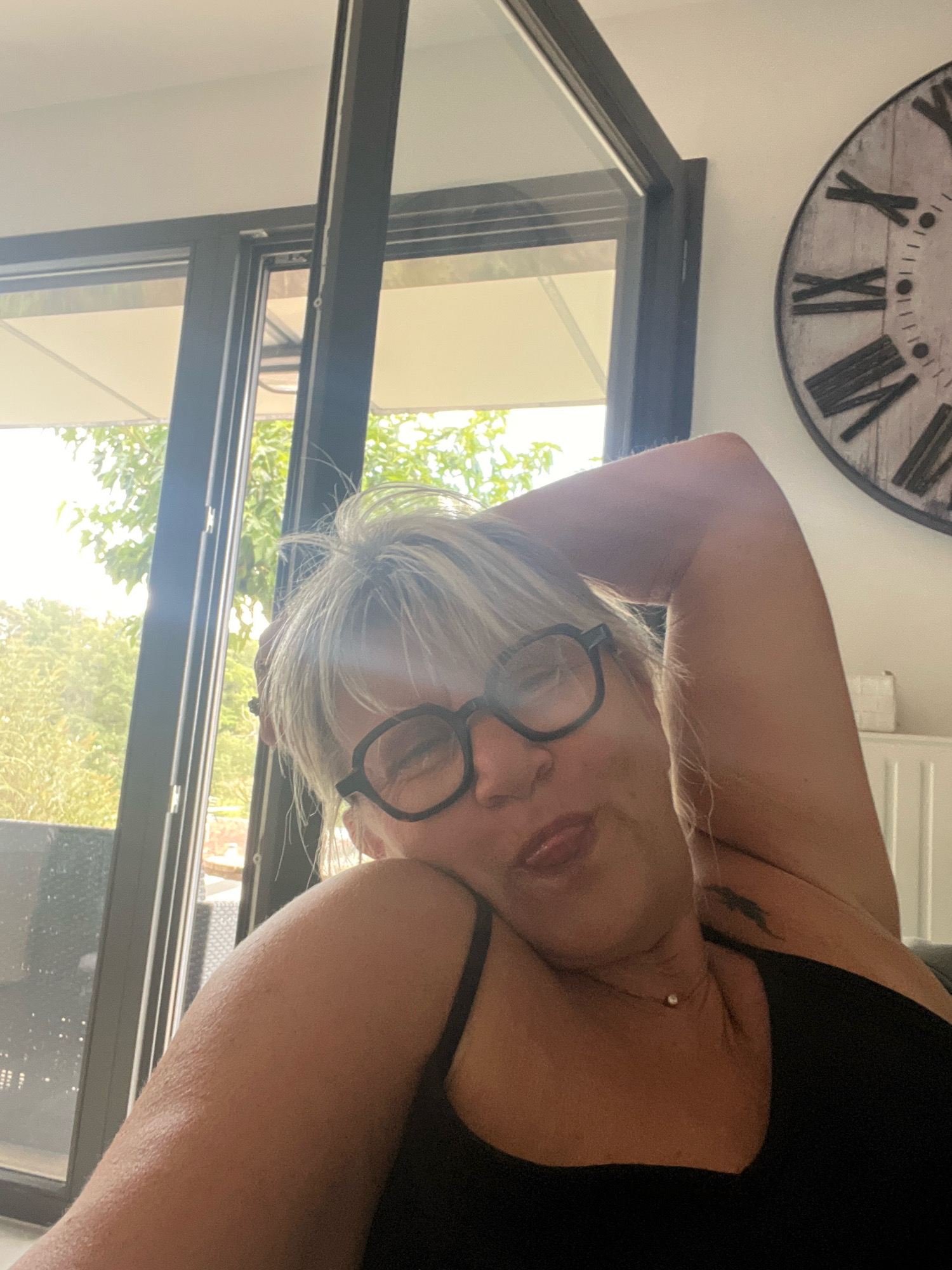 The image shows a woman with blonde hair, wearing glasses and a black tank top. She is leaning back with her arm behind her head, and her tongue is sticking out slightly. She is wearing a delicate necklace with a single pearl. The background shows a large window with trees visible outside, and a clock on the wall. The location is identified as Chambœuf, France.