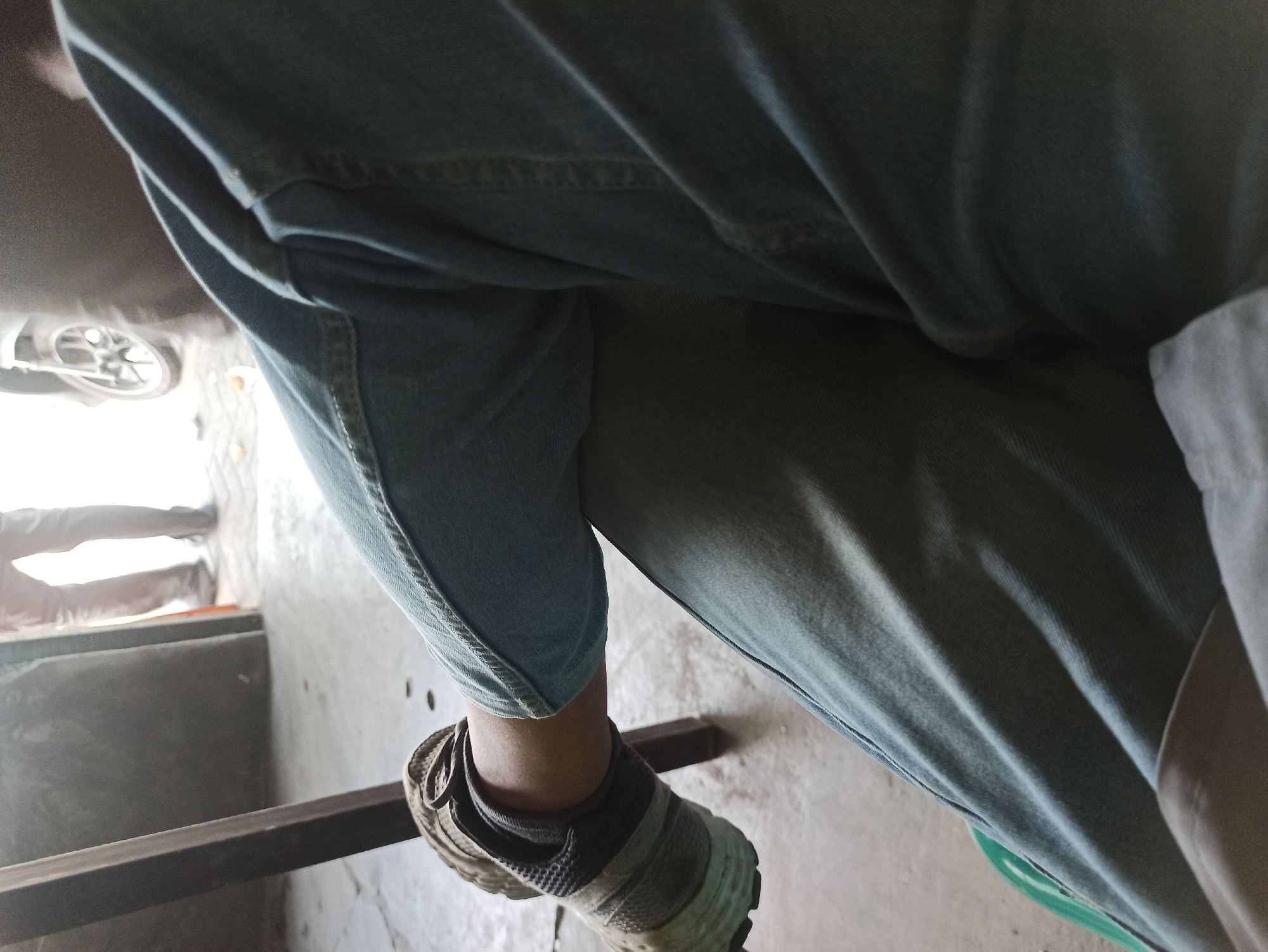 An individual's legs, clad in light blue denim jeans, are prominently featured in the foreground of this image. The person is wearing a dark and beige athletic shoe on their left foot, which rests near a dark vertical object, possibly a table leg or chair. The background suggests an indoor or semi-outdoor setting with a concrete floor. In the middle ground, another individual is partially visible, standing and facing away from the camera, their lower legs also covered in light-colored pants. Further in the background, the wheel of a motorcycle is discernible, indicating a public or roadside environment. The lighting suggests it is daytime. This scene likely depicts a casual moment, possibly at a roadside eatery or resting spot in Medan, Indonesia.