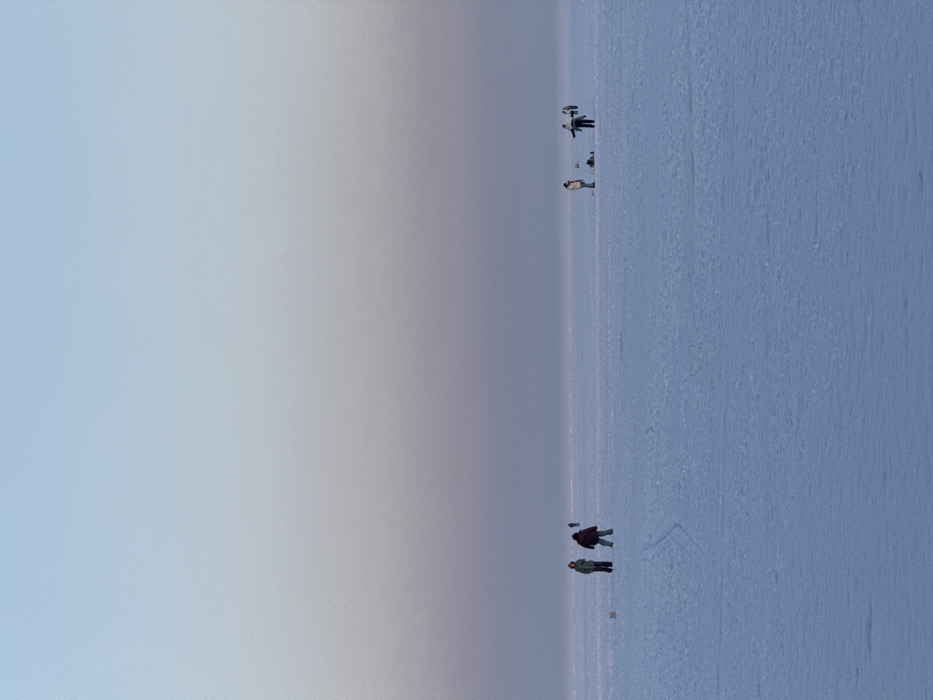 The image displays a vast, flat, snow-covered landscape, characteristic of a frozen body of water, situated in Liutizh, Ukraine. The sky transitions from a pale blue overhead to a muted purple-grey at the distant horizon, indicating ambient light conditions. Six human figures are observed across the mid-ground.
On the left, two individuals stand on the frozen surface. One wears a light-colored jacket and dark pants, facing slightly right. The second, in a dark reddish jacket and dark pants, faces forward. A small, light-colored object rests on the snow to their left. Faint linear impressions are visible on the snow behind them.
On the right, a group of three individuals is clustered. The leftmost wears a light jacket and dark pants, facing left, with a dark object on the snow nearby. A central figure, in a dark jacket and dark pants, stands with arms extended horizontally. A third person, in a light jacket, is positioned further back and to the right within this cluster. A sixth, indistinct figure appears as a dark silhouette further right on the horizon line.
The individuals are spaced out across the expansive, uniform frozen surface. Their primary action consists of standing. Specific fishing gear or active fishing is not clearly discernible, though the presence of objects on the ice near the groups could be consistent with ice fishing equipment.