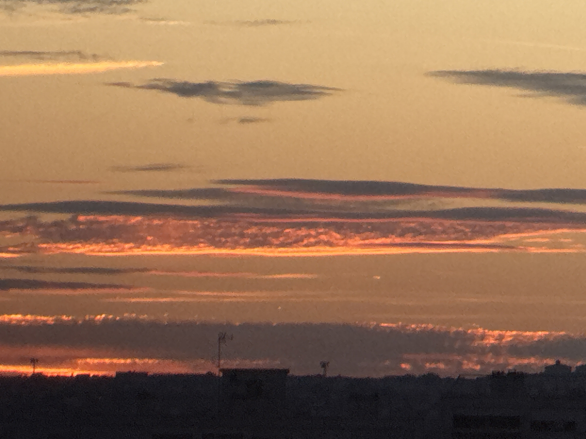 The image shows a dusky sky over a silhouetted cityscape. The sky is a gradient of warm oranges and yellows, with distinct horizontal cloud formations. Some clouds are illuminated from below with fiery orange and red hues, while others are darker grey. In the lower portion of the image, the dark silhouettes of buildings and structures are visible, with a few faint lights suggesting urban activity. A tall, thin antenna or pole is visible on the left side of the silhouette. The overall impression is of a sunset or twilight over an urban area.
