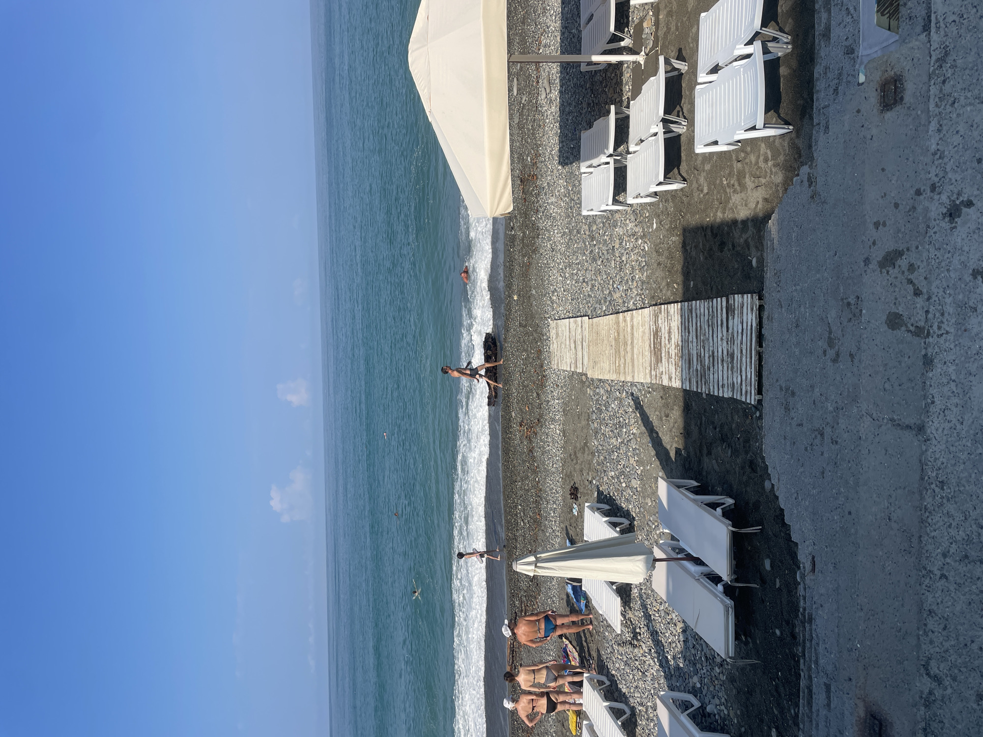 The image depicts a pebble beach scene on a sunny day, likely in Sochi, Russia, given the typical resort atmosphere. The turquoise water of the sea is visible in the background with gentle waves and a few swimmers. In the foreground, rows of empty white sun loungers and a large beige umbrella suggest a beach resort area. A wooden boardwalk leads from the upper portion of the scene towards the water. Several individuals are present on the beach: a man is walking towards the water, a group of four people (two men and two women) are standing near the water's edge, and two other individuals are swimming. The beach itself is composed of dark grey pebbles. The sky is a clear blue with a few wispy white clouds.