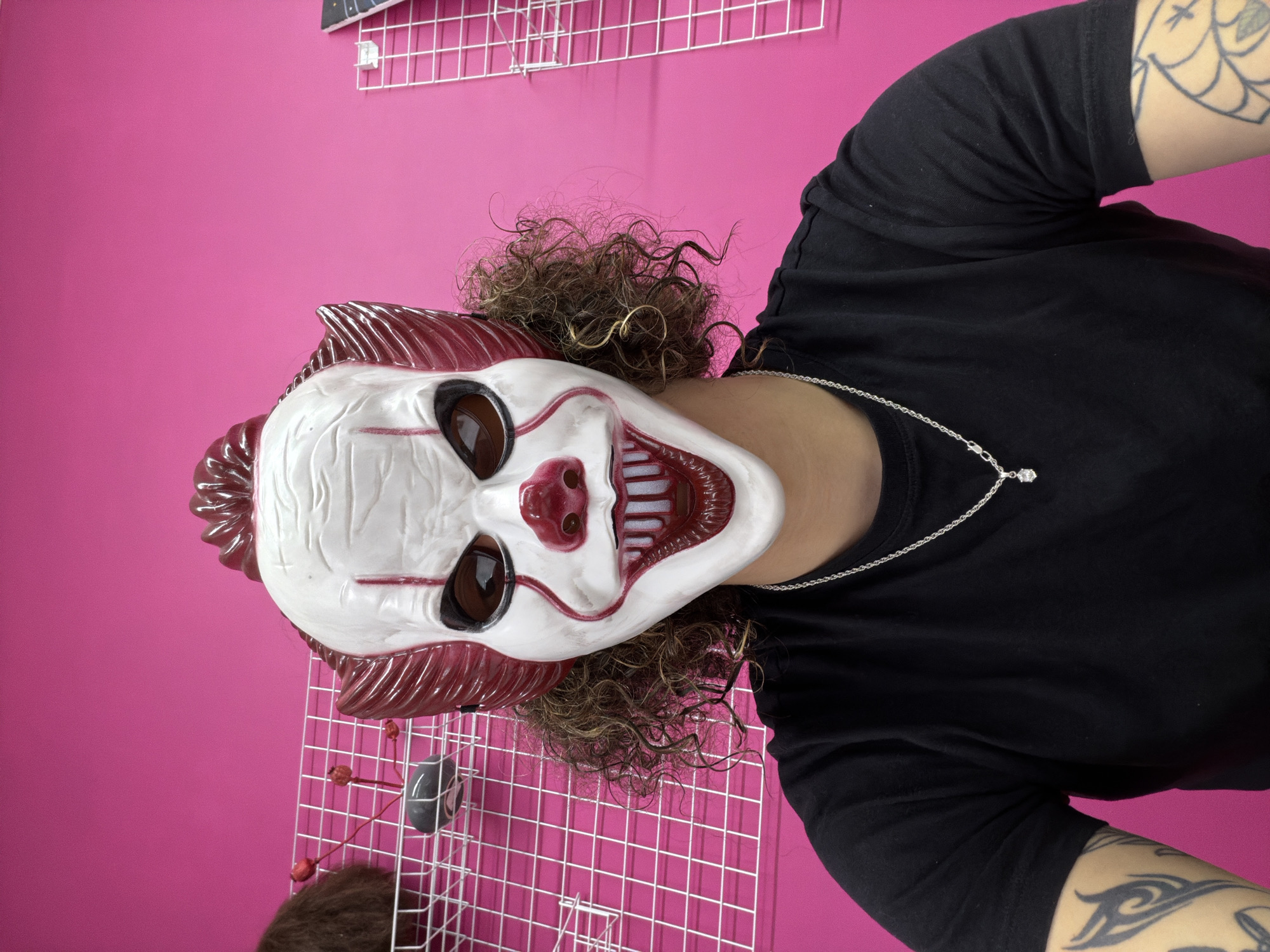 A person wearing a white and red clown mask with dark eye sockets, a red nose, and red lips smiles widely at the camera. The mask has red hair on the sides and a red pompadour on top. The person's curly brown hair peeks out from beneath the mask. They are wearing a black t-shirt with a silver chain necklace that has a small diamond pendant. The background is a bright pink wall with a white grid shelf mounted on it. On the shelf, there are various items, including what appears to be a brown furry object and a small grey and black object. The scene is indoors, in a brightly lit room.