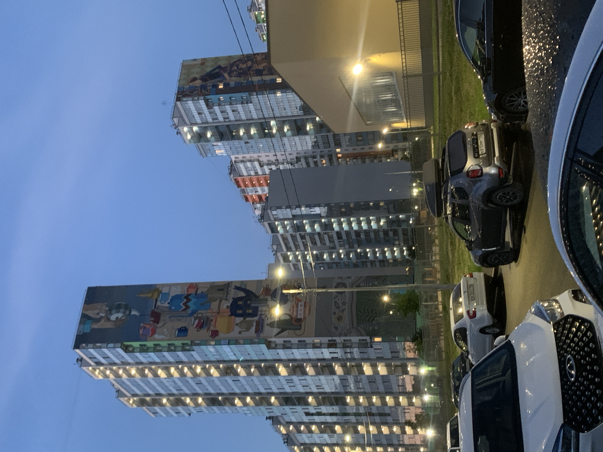 This media file depicts an urban scene at dusk in Saint Petersburg, Russia. The primary subjects are two multi-story apartment buildings adorned with large murals. The building on the left features a mural with a cartoonish, childlike style, depicting various figures and objects, possibly a beach scene. The building on the right has a mural that is less clear but appears to be an abstract or patterned design.
In the foreground, several cars are parked, suggesting a residential area. These vehicles include a silver SUV with a roof box, a black car, and parts of white cars. The ground appears wet, reflecting the artificial lights, indicating recent rain or damp conditions. The sky is a deep blue, typical of twilight, with artificial lights from the buildings and streetlights casting a warm glow.
There is no visible activity or event taking place, beyond the general ambiance of an evening in a populated urban environment. Notable details include the time of day (dusk/early evening), the weather (damp, likely after rain), and the location cues of apartment buildings with murals, characteristic of some modern Russian urban developments. The overall mood is calm and slightly subdued, influenced by the fading light and the presence of the parked cars. There is some Cyrillic text visible on the license plate of the silver SUV.