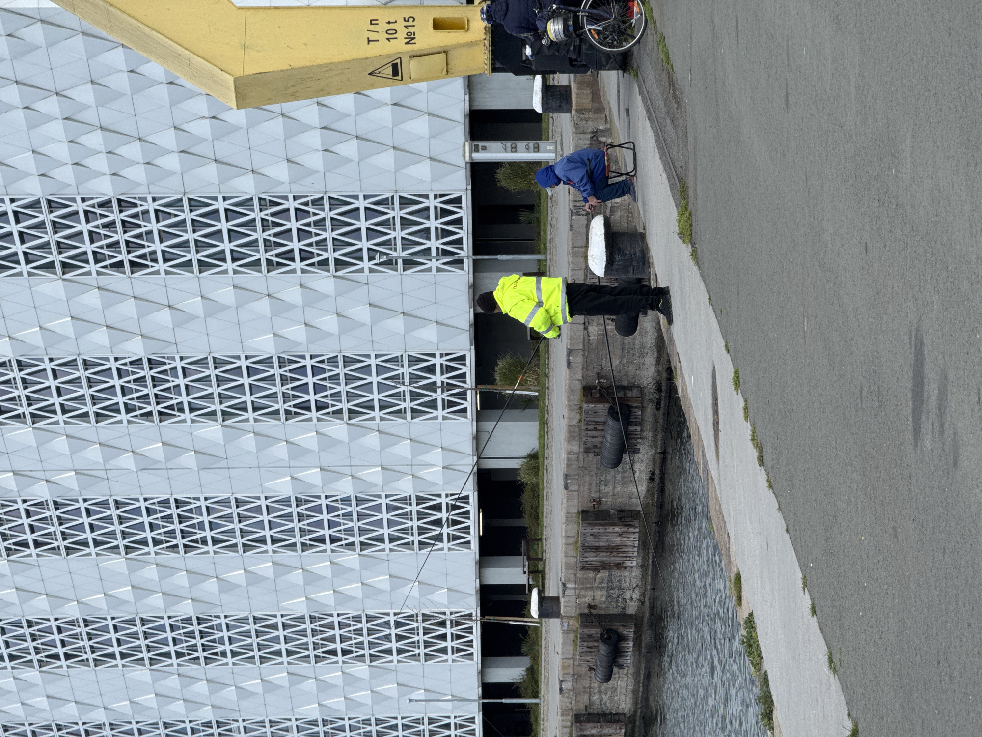 The image shows a quay in Burgas, Bulgaria. Two people are fishing: one wearing a yellow jacket standing, and another wearing a blue jacket sitting on a small stool. A bicycle is parked behind the seated fisherman, next to a yellow industrial element marked "T/n 10 t No 15." The quay’s edge consists of concrete and wooden elements. A modern building with a geometric patterned facade is behind the people. The water is to the left of the image, and a road is to the right.