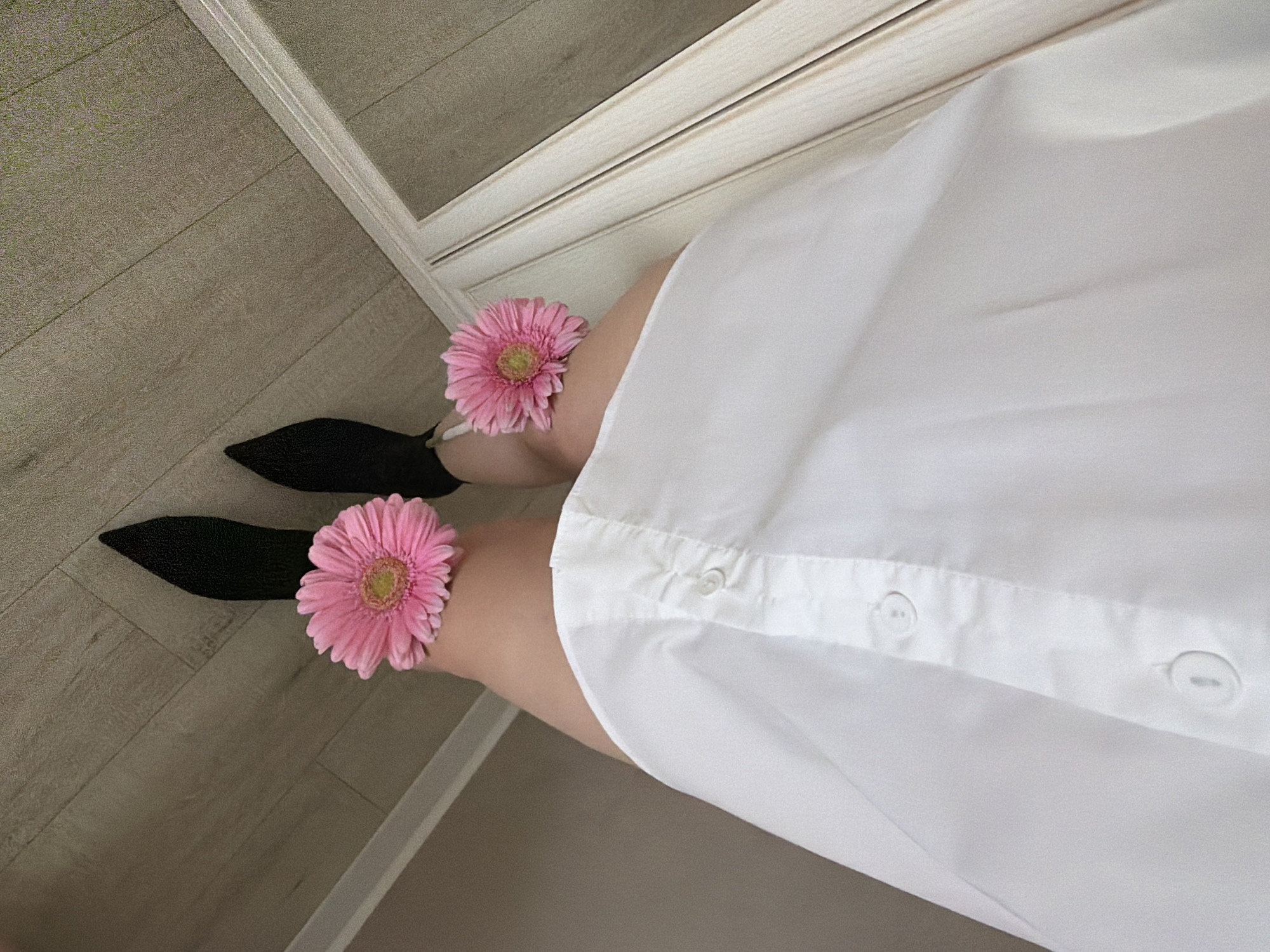 A high-angle shot depicts a person wearing black shoes, a white dress shirt, and adorned with two pink Gerbera daisies placed at the knee level. The scene is set indoors, next to a wall with white trim. A floor with a wood-grain pattern is visible. There is no information to suggest the location is in Votkinsk, Russia.