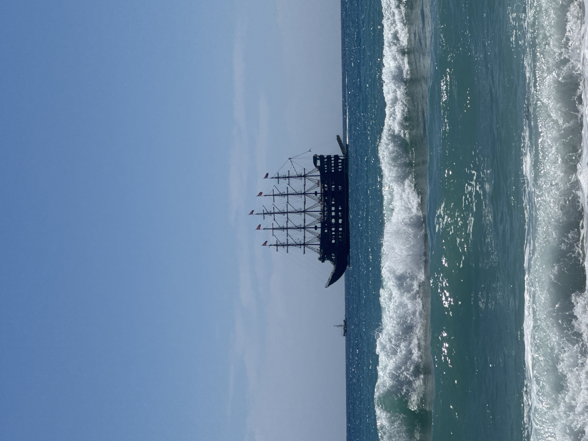 The image shows a tall sailing ship with multiple masts on the Mediterranean Sea in Alanya, Turkey. The ship is dark in color with red flags on the masts. The sky is clear blue with a slight gradient from light to dark towards the horizon. The sea is deep blue with white waves breaking along the shoreline. Another smaller boat is visible further in the distance.