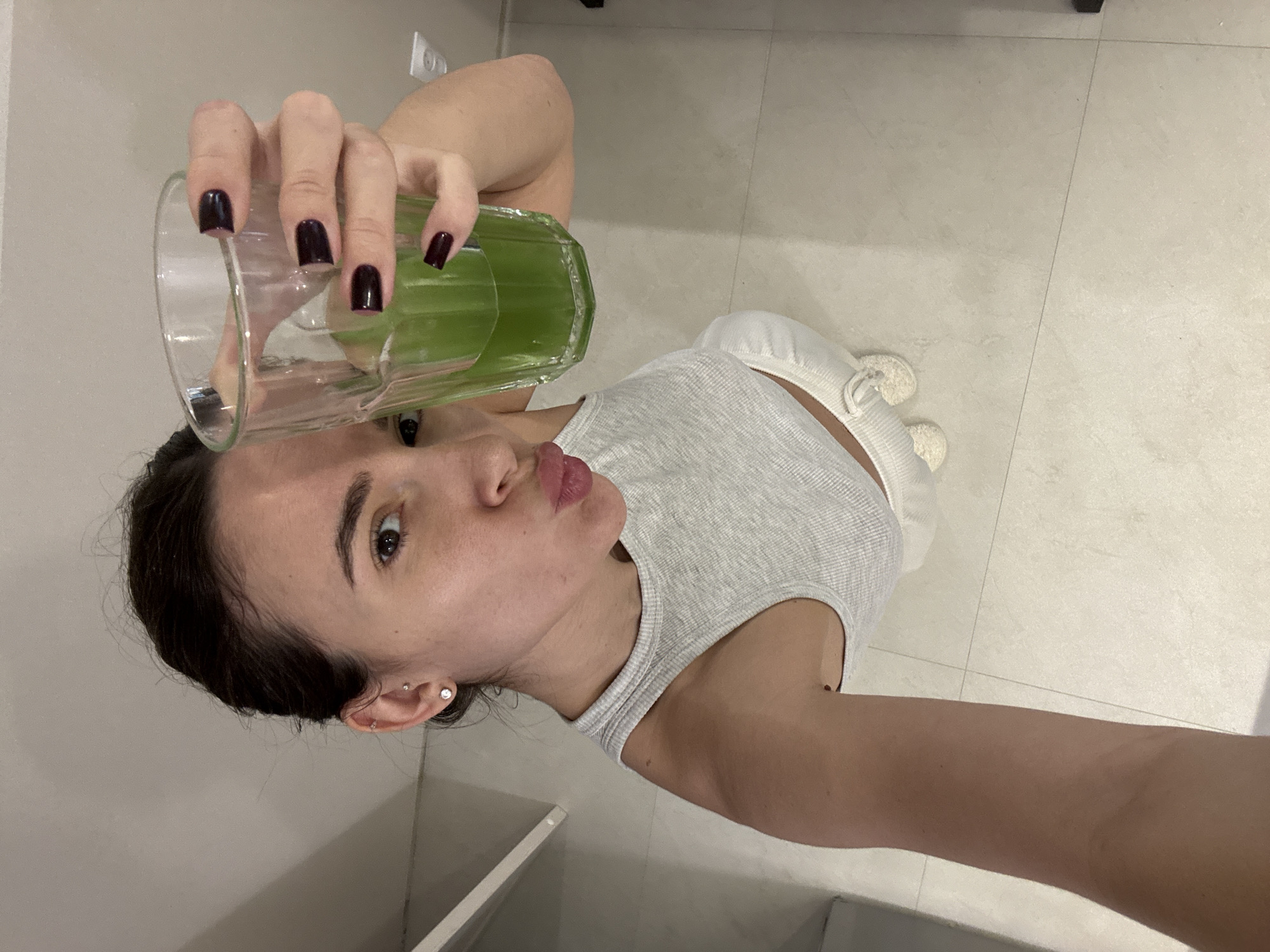A woman is holding up a glass of green liquid, possibly a smoothie or juice. She is wearing a gray tank top and white bottoms, and has dark hair pulled back. She has dark nail polish and several earrings in her left ear. The background appears to be a room with tile flooring and a white wall and possibly white cabinetry in the lower left corner. There is no information in the image to indicate this is Chaikovsky, Russia.