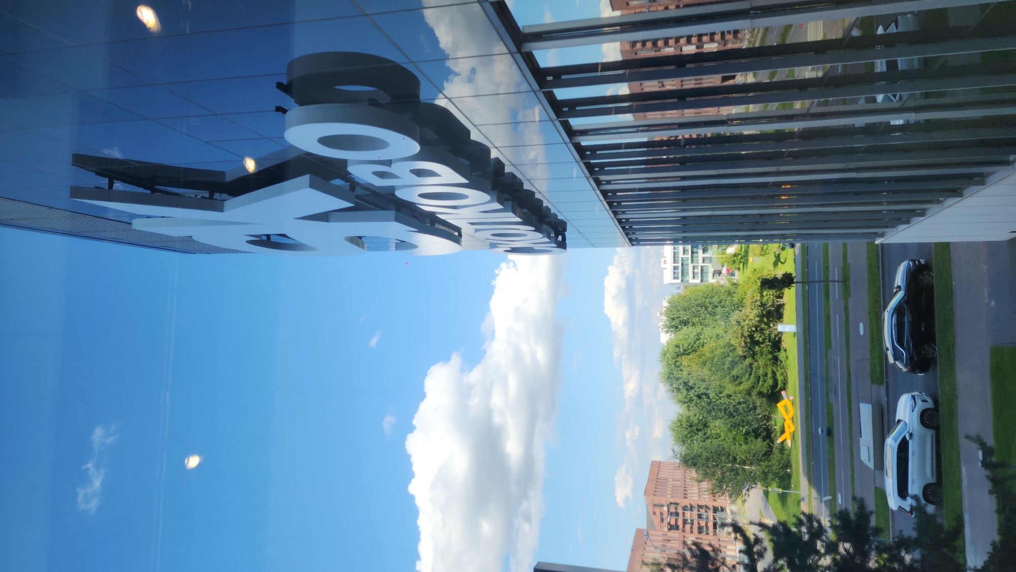 The image captures a view through a window of a modern building with a glass facade. The building prominently features large, white, three-dimensional lettering that reads "SKOLKOVO". The facade also reflects the blue sky with scattered white clouds and other buildings.
In the background, a tree-lined street is visible with several buildings, including one with a reddish-brown brick facade and another with a white, modern design. There is a yellow sculpture of an airplane on a grassy area between the road and the buildings. Two cars, a white SUV and a black SUV, are parked on the side of the road. The overall scene suggests an urban or suburban environment, likely a business or innovation district.
Based on the prominent "SKOLKOVO" lettering, the location is likely within the Skolkovo Innovation Center, a technology park located in the Odintsovsky District of Moscow Oblast, Russia, just west of the Moscow city limits.