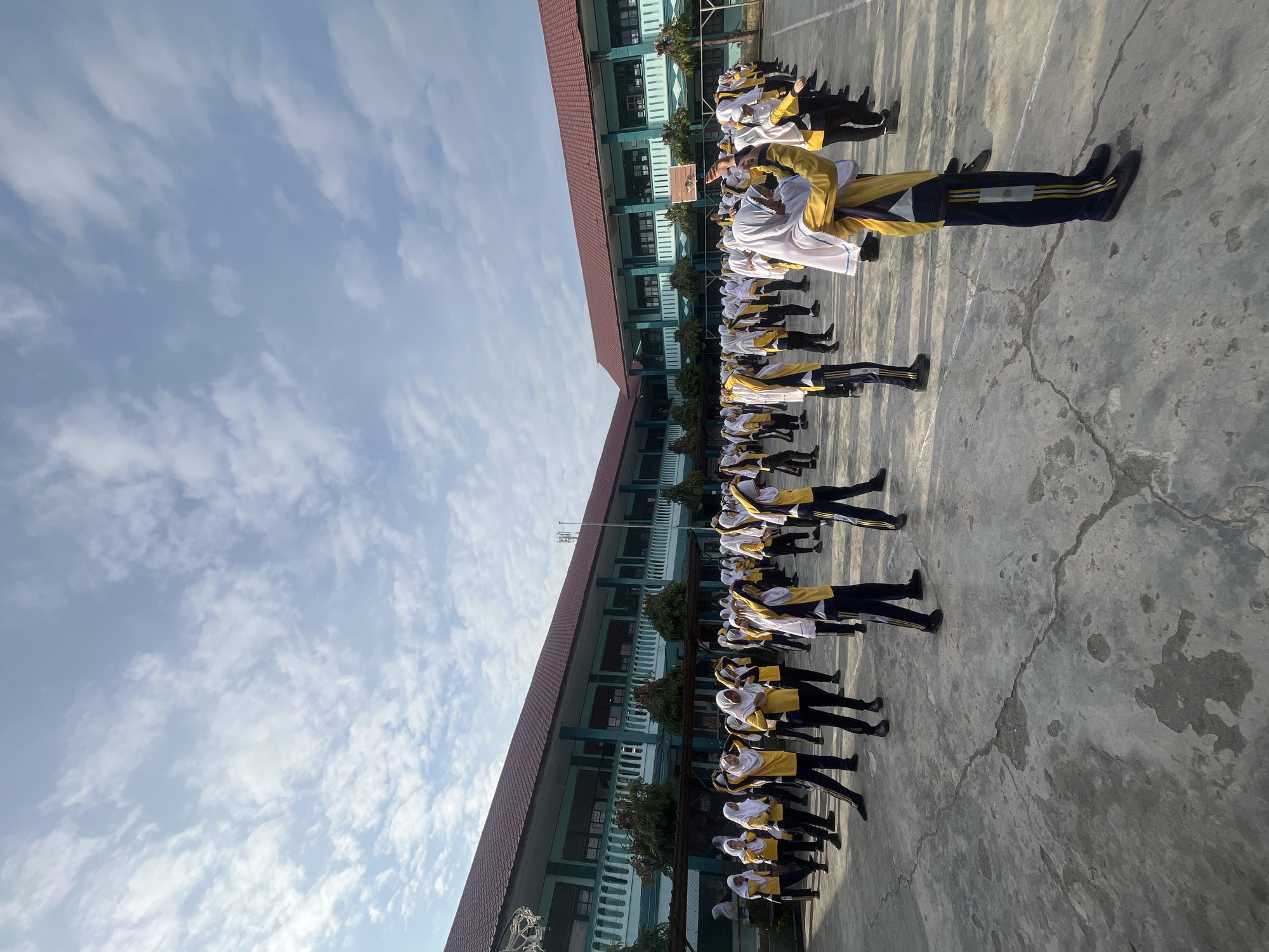 A group of students, primarily female, wearing yellow shirts, dark pants, and white headscarves, are participating in an outdoor physical activity, possibly exercise or a school assembly. They are lined up in rows on a paved courtyard in front of a two-story school building. The building has a red-tiled roof and teal-colored walls with white trim. Trees and a basketball hoop are visible around the courtyard. The sky is partly cloudy and appears to be early morning or late afternoon. The students are performing synchronized arm movements. The scene is set in the city of Lhokseumawe, Indonesia.