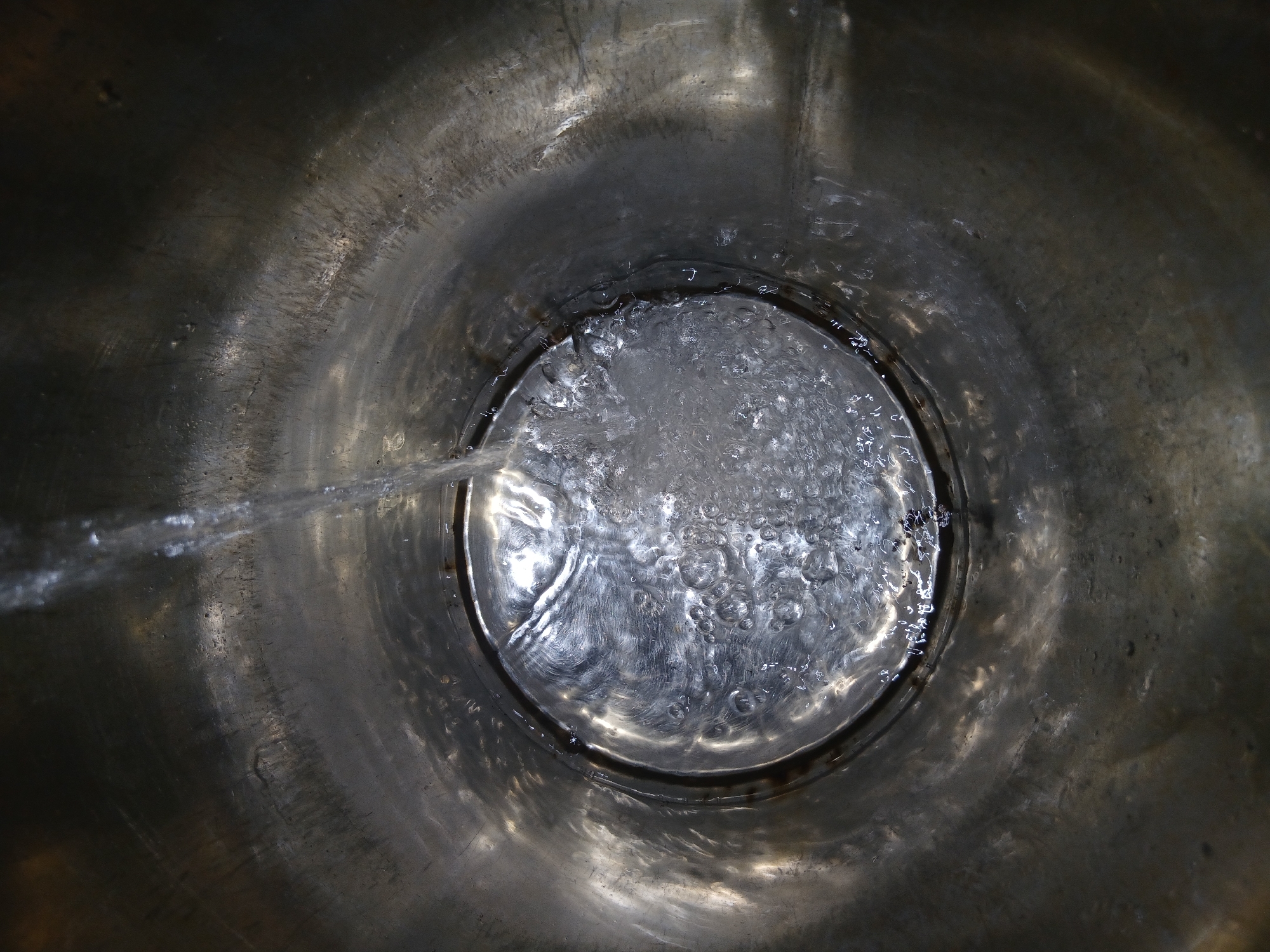 The image shows a top-down view into a metal container, likely a bucket or tub, partially filled with water. Water is being poured into the container from the left side, creating ripples and bubbles on the water's surface. The metal of the container appears worn and has a grainy texture. The location is unknown and cannot be determined from the image.