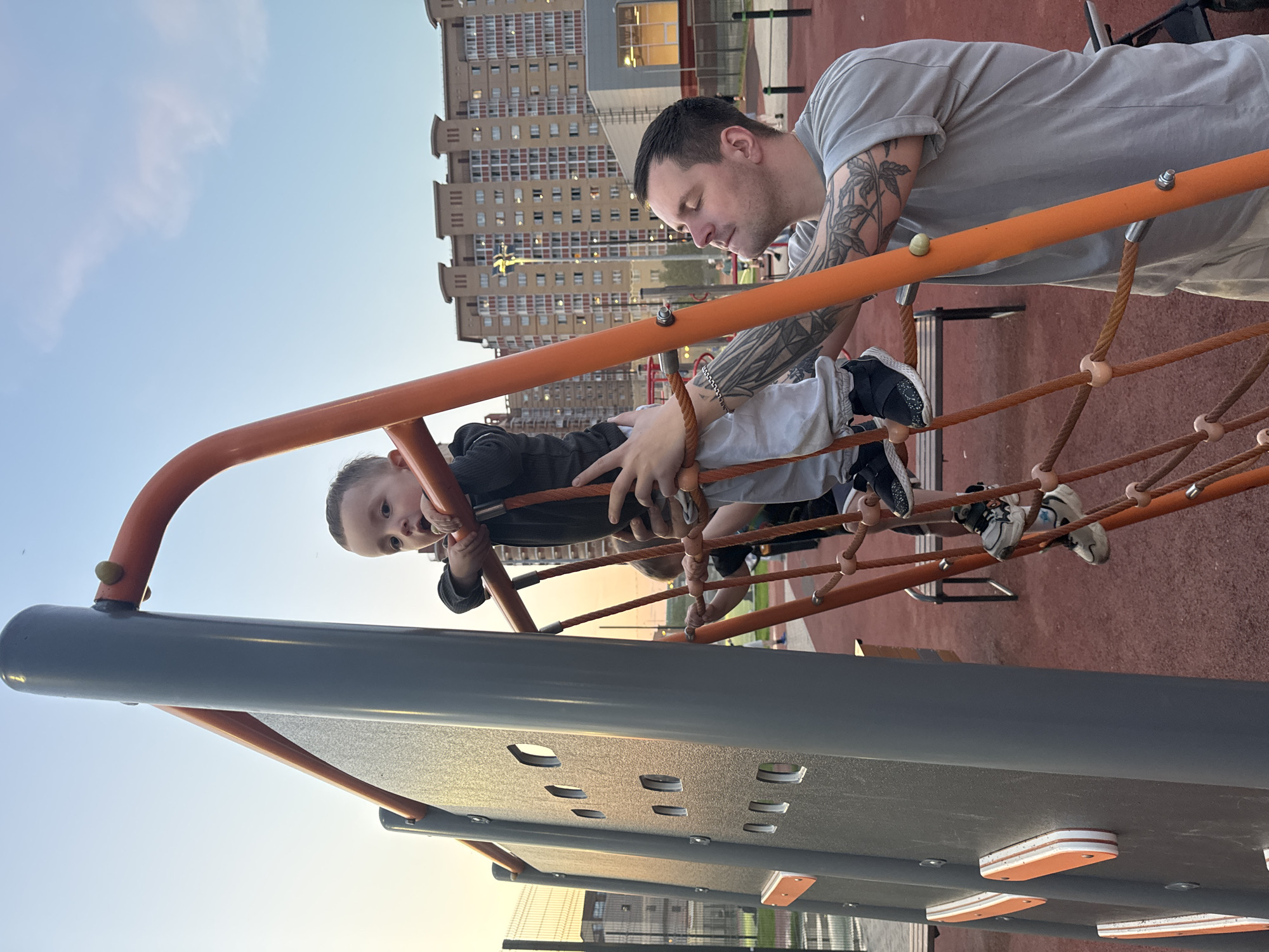 The image shows a man and a young child on a playground structure. The man, with tattoos on his arms, is wearing a gray t-shirt and is helping the child navigate a rope climbing net. The child is wearing a dark jacket and light-colored pants with dark shoes. The structure appears to be a climbing frame with orange metal supports and brown rope netting. The ground below is a reddish-brown color, possibly a safety surface. In the background, there are tall apartment buildings, suggesting an urban environment. The image appears to be taken in Vniissok, Russia due to the building style.