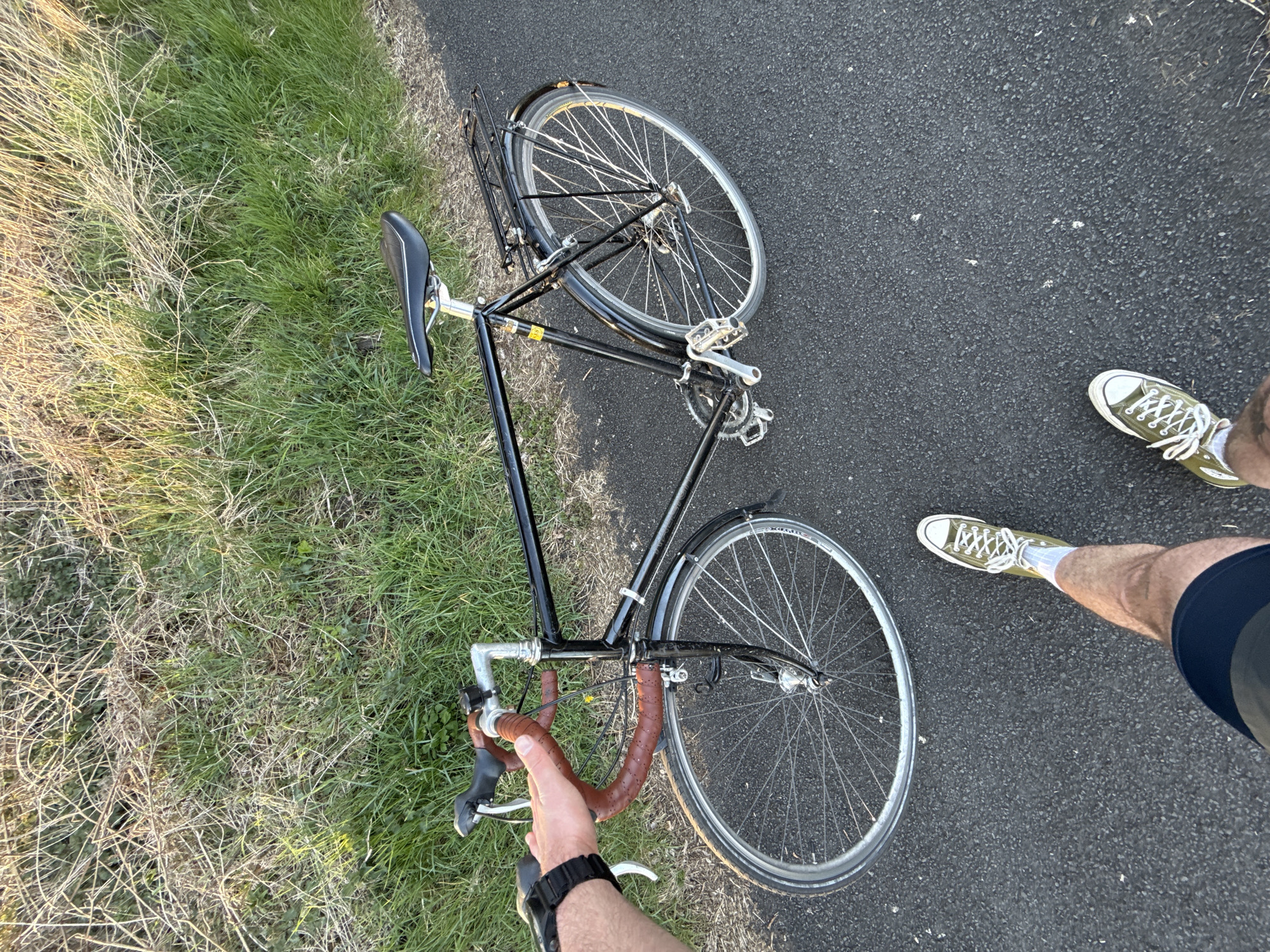 The image captures a first-person perspective of a person standing next to a black bicycle on an asphalt path. The photo is rotated, showing the scene diagonally.
The primary subject is a classic-style road bicycle with a black frame, silver wheels, fenders, and a rear rack. It features brown handlebar tape and a black saddle. The bicycle appears to be resting on the ground or being held by the person, who is visible from the waist down, holding the handlebars with their right hand. A black digital watch is visible on the person's wrist.
The person is wearing dark shorts and olive green/khaki canvas sneakers with white laces. Their left leg and foot are visible on the asphalt to the right of the bicycle.
The setting is outdoors, with a dark gray asphalt path on one side and a grassy area on the other. The grass transitions from vibrant green near the path to drier, lighter-colored stalks further away. Sunlight casts long shadows and highlights the textures of the grass, suggesting it is daytime, possibly late afternoon or early morning. No specific location cues are identifiable beyond an outdoor, possibly rural or park-like environment. There is a small yellow sticker on the bicycle frame, but its text is illegible.