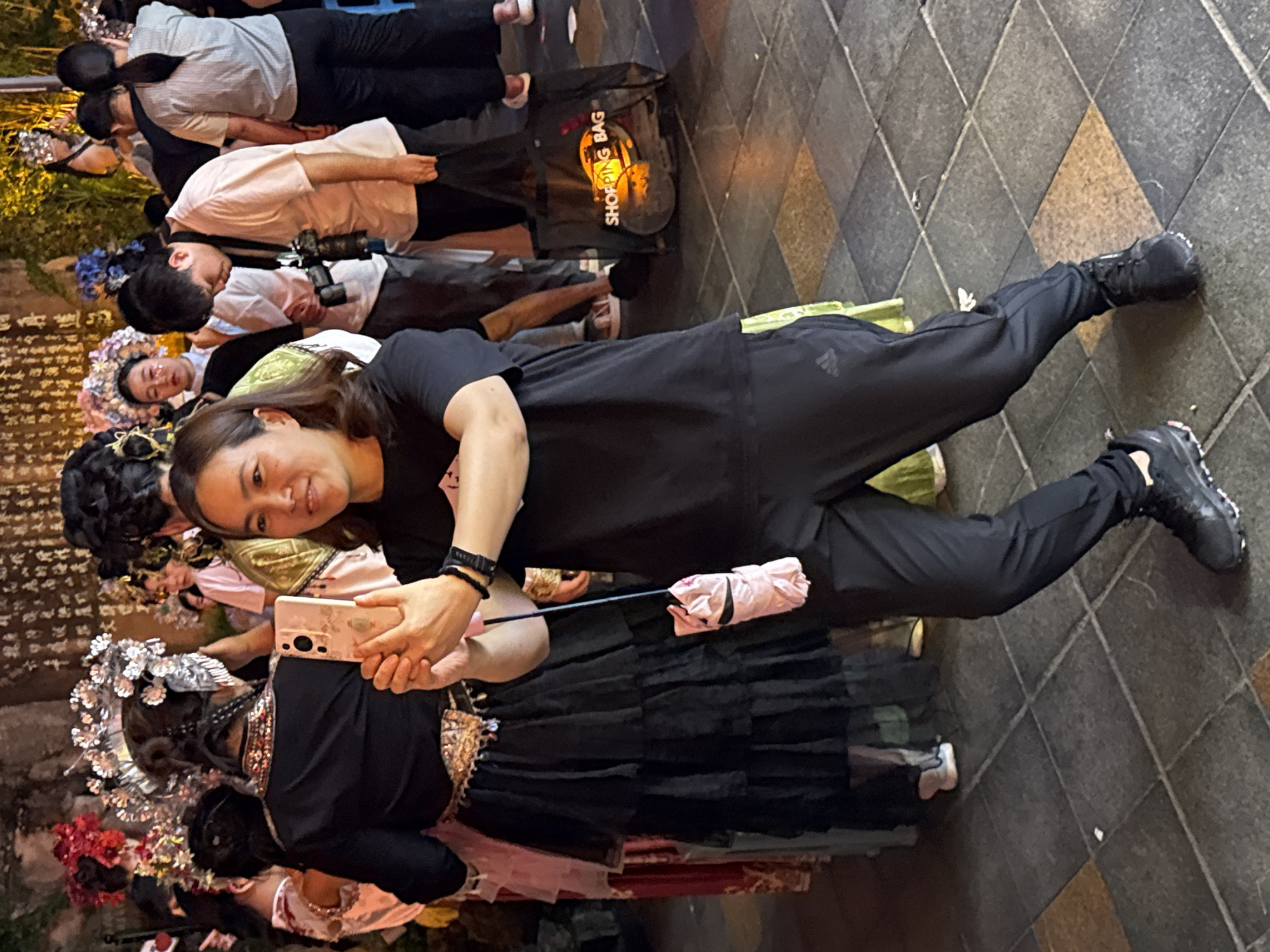A woman in athletic wear stands on a tiled surface, holding a smartphone in her right hand to take a photo of people dressed in traditional costumes. She is wearing a black t-shirt and black pants with an Adidas logo visible on the left pant leg. She has dark hair and is smiling. To her left, a woman in a black tiered dress and an elaborate headdress is visible, also facing away from the camera. Other individuals in various costumes and modern attire are present in the background. A pink, folded umbrella hangs from a string near the woman taking the photo. In the background, a black shopping bag with "SHOPPING BAG" printed on it is visible. The scene appears to be outdoors in an urban setting, possibly during an event or festival in Yuzhong District, China, indicated by the tiled ground and architectural elements in the background.