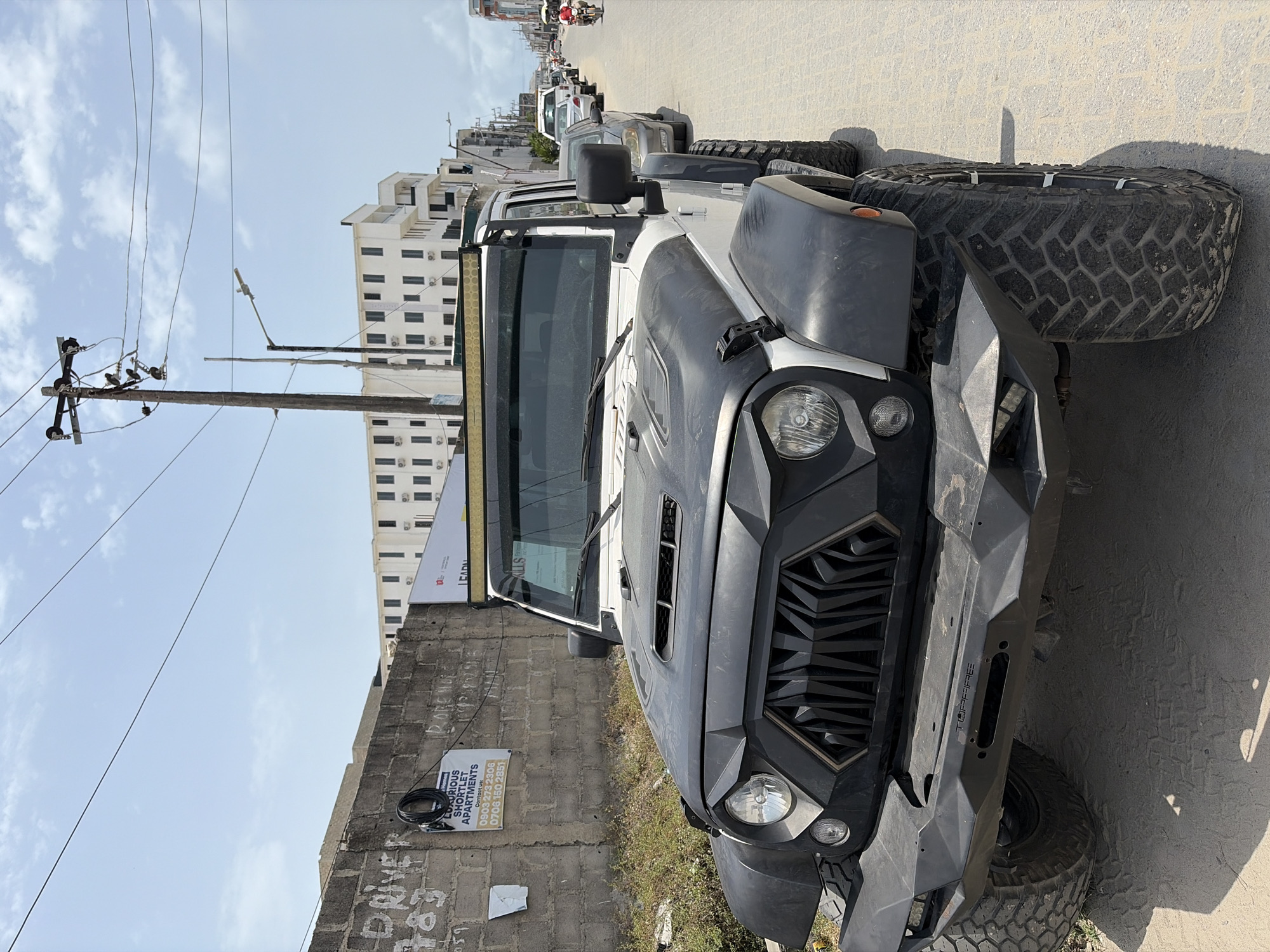 The image shows a heavily modified Jeep Wrangler parked on the side of a street in what appears to be Igboefon, Nigeria. The Jeep has a black and white color scheme, oversized tires, an aftermarket grille, and a robust front bumper with the word "TOPFREE" visible on its side. The vehicle is parked next to a low brick wall covered in graffiti and advertisements, including one for "Luxurious Shortlet Apartments" with a phone number. In the background, there are buildings, utility poles with power lines, and other vehicles parked along the street. In the far distance, there are hints of people and traffic, suggesting a relatively busy urban environment. The sky is mostly clear with some clouds.