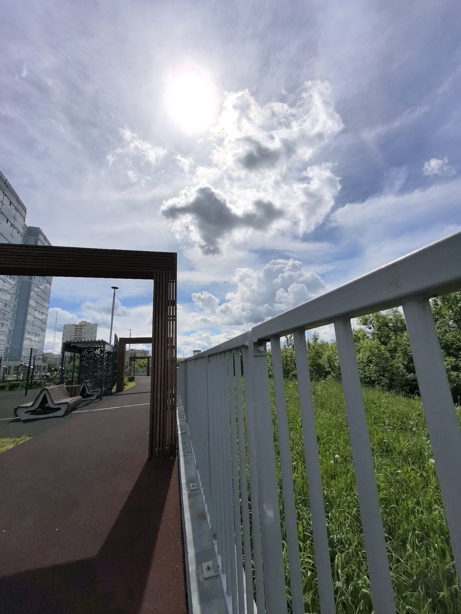 The image shows a perspective from a raised walkway or bridge in Perm, Russia. To the left is a park area with benches and a modern structure, possibly for exercise or recreation. Beyond this park area, a tall building is visible. A brown wooden frame structure stands near the edge of the walkway. To the right, a grey metal railing runs along the walkway's edge, separating it from a patch of tall green grass and trees. The sky is partly cloudy with the sun shining brightly through the clouds.
