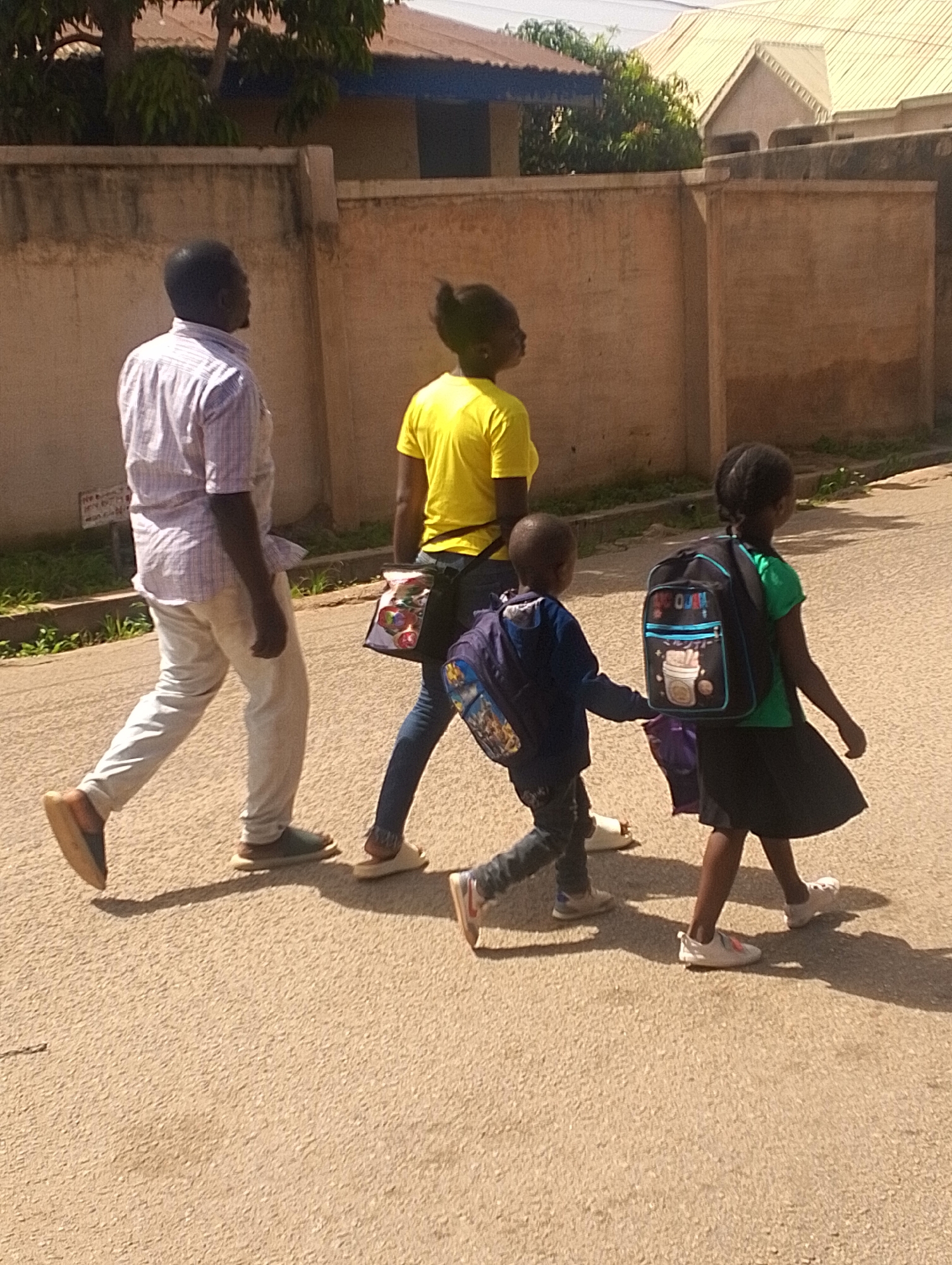 In this medium-shot outdoor photograph, three individuals are captured in motion, walking on a paved road. The scene appears to be a sunny day, likely in the morning or afternoon given the strong shadows cast by the subjects. The background features a weathered, tan-colored wall that runs horizontally across the frame, with glimpses of buildings and trees behind it.
The leftmost subject is an adult male, seen from the knees up, wearing a light-colored, checkered short-sleeved shirt and light-colored trousers. He has dark skin and is walking away from the camera, his body angled to the right.
To his right, a young woman or older girl is walking, also facing away from the camera and angled slightly to the right. She is wearing a bright yellow t-shirt and dark trousers. A colorful, patterned bag is slung over her shoulder.
In front of the woman, two children are walking, one slightly ahead of the other. The child on the left is a boy, wearing a dark blue jacket and dark trousers. He is carrying a blue backpack with some images on it, and appears to be holding the hand of the child to his right. The child on the right is a girl, wearing a green t-shirt and a dark skirt. She is also carrying a backpack, which is predominantly black with colorful designs and text that reads "OOH!". Both children are wearing school-appropriate shoes.
The overall atmosphere suggests a casual outing, possibly children walking to or from school, accompanied by an adult. The lighting indicates a bright day, and the paved road and wall are common elements in urban or semi-urban environments. No specific emotions are discernible from the subjects' postures, but their forward movement suggests a purposeful journey. The location context is Jos, Nigeria, which aligns with the warm weather suggested by the bright sunlight and shadows.