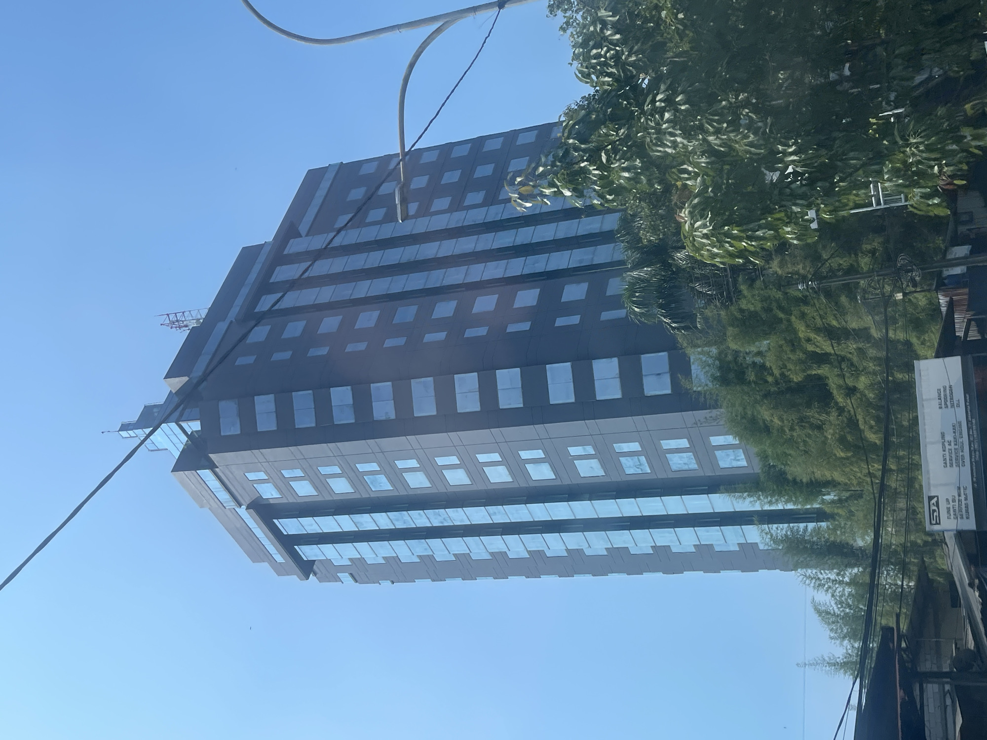 A tall, modern skyscraper with a dark facade and blue glass windows stands against a clear blue sky in Central Jakarta, Indonesia. The building has a stepped roofline and a small antenna array at its summit. In the foreground, lush green trees, including bamboo, partially obscure the lower floors of the building. A street lamp with a curved pole and wires is visible to the right. At the bottom of the image, a sign with Indonesian text and a logo is partially visible, suggesting a commercial or service area. The overall scene depicts an urban environment with a mix of modern architecture and natural elements.