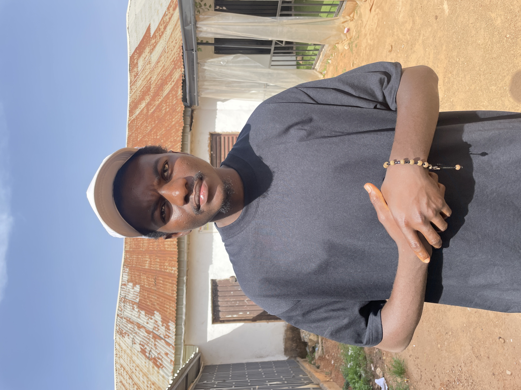 The image features a man with dark skin, a short beard, and a tan bucket hat standing outside. He is wearing a dark gray t-shirt and has a beaded bracelet on his left wrist. He is standing in front of a one-story building. The building is painted white with a rusty corrugated metal roof. A barred window is visible on the side of the building. The ground is a mixture of dirt and vegetation, suggesting a residential area. The photograph was taken during daylight hours under bright sunlight. There is no precise information that indicates that the image was taken in Barakin Akawo, Nigeria.