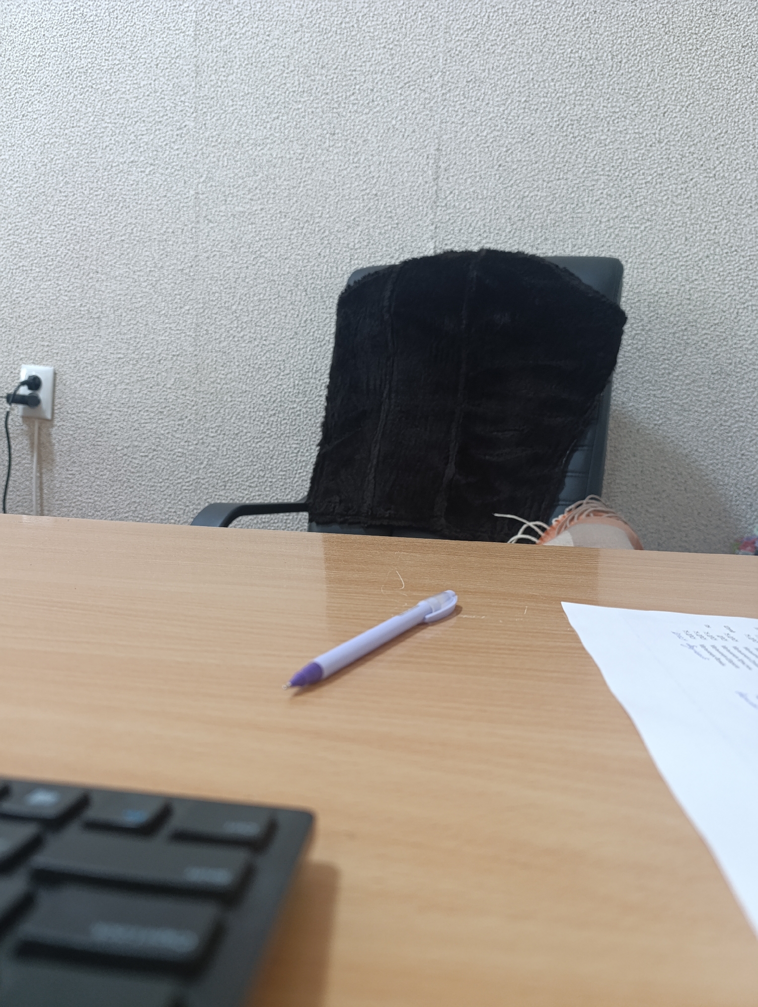 An eye-level, close-up shot shows a light wood desk with a black keyboard in the lower left, and a white sheet of paper and a purple pen with a metal tip on the right. In the background, a black office chair with a fuzzy black cover is visible against a textured white wall with a power outlet.