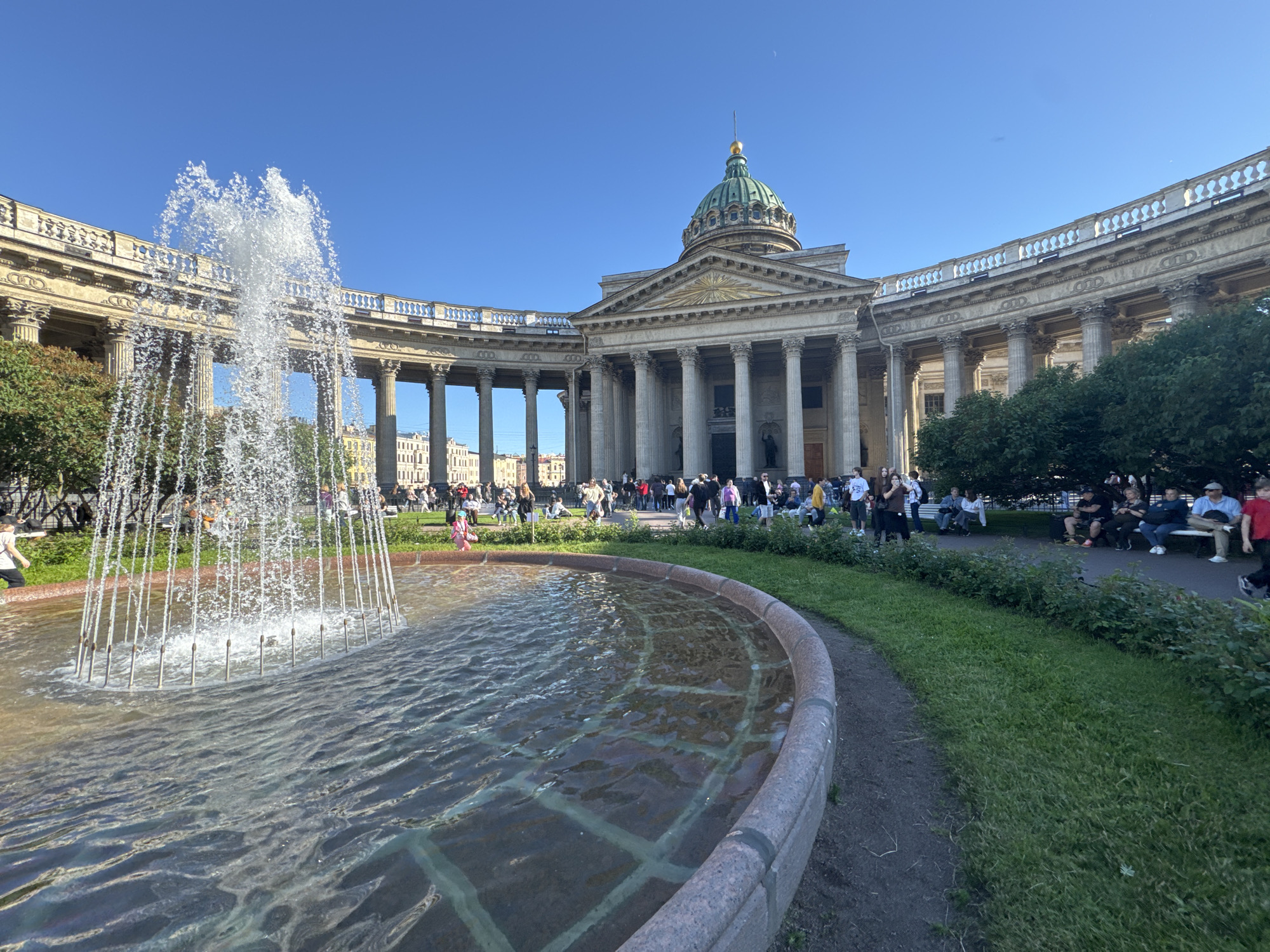 This is a photograph of Kazan Cathedral in Saint Petersburg, Russia, on a clear, sunny day. In the foreground, a large, circular fountain with stone edging is active, with water spraying upwards. The water in the fountain basin has a textured, rippled surface, and a grid pattern is visible beneath the water. A paved pathway runs alongside the fountain.
Behind the fountain, the grand colonnade of Kazan Cathedral curves gracefully. The colonnade is made of stone, with numerous large columns supporting an ornate upper structure. Beyond the colonnade, the central dome of the cathedral is visible, topped with a golden spire. The sky is a vibrant, cloudless blue.
A crowd of people is dispersed throughout the scene, some walking along the path or the grass, others sitting on benches or in front of the cathedral. The people are dressed in casual, warm-weather clothing. Trees and green grass are visible to the right of the colonnade, with more people gathered in this area. The architecture of the cathedral is classical and imposing, with a sense of grandeur. The overall atmosphere is one of a public gathering place on a pleasant day, with the iconic cathedral as the focal point.