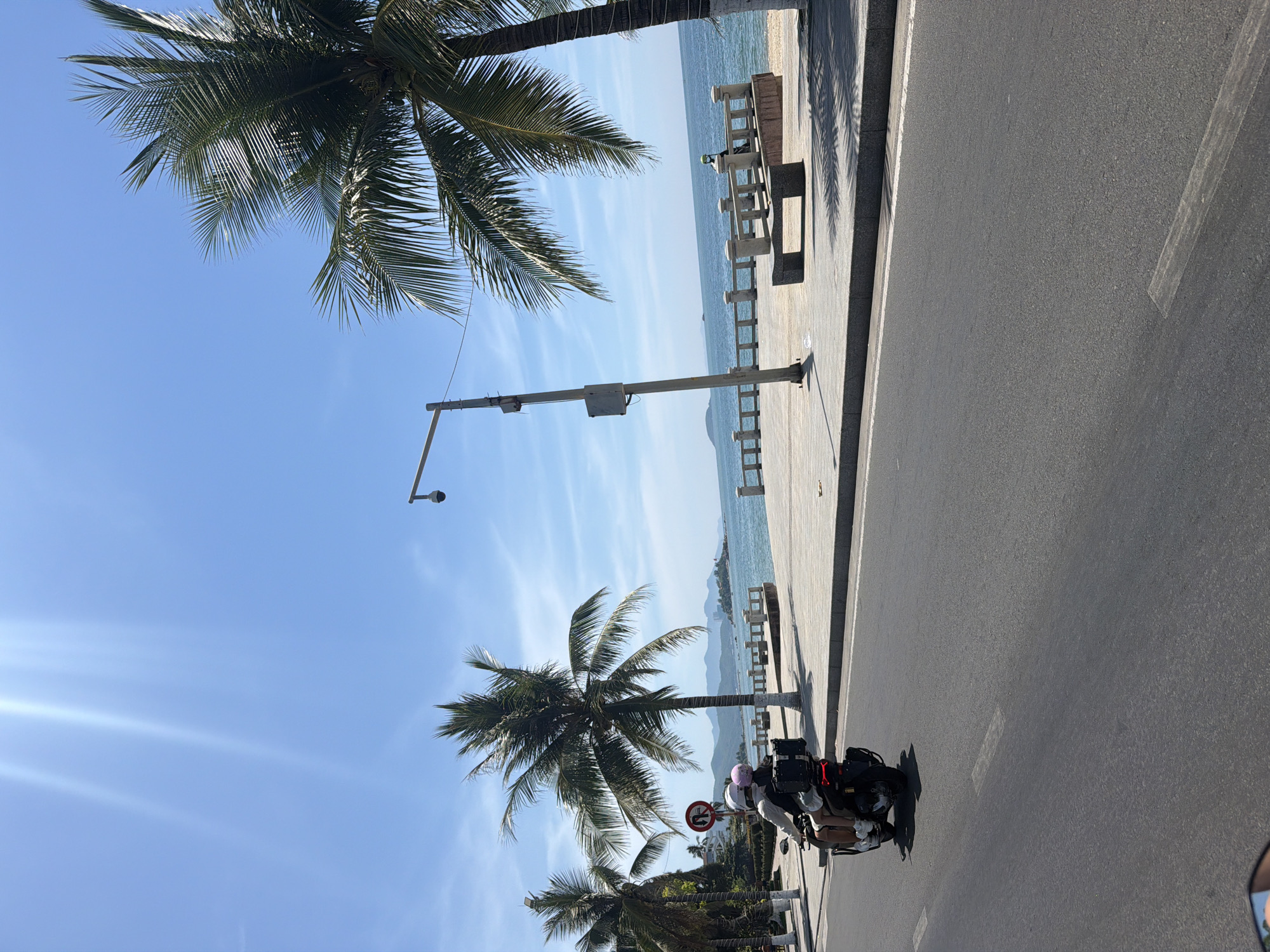 The image captures a bright, sunny day with a clear blue sky over the coastal city of Nha Trang, Vietnam. In the foreground, a motorcycle with two passengers, a man and a woman wearing helmets, travels along an asphalt road. The woman is seated behind the man and is wearing a white top and dark shorts, while the man is wearing a light-colored shirt and shorts. They are both wearing helmets, one white and the other pink. A signpost with a circular red and white sign is visible to the left of the road.
Beyond the road, a paved walkway lines the ocean. Palm trees with lush green fronds dot the landscape, some leaning over the walkway. A lamppost with a security camera is mounted on the walkway. A stone bench sits on the walkway, casting a shadow from the palm trees. The turquoise ocean stretches to the horizon, where faint outlines of islands or mountains can be seen. The water is calm, with gentle waves lapping at a sandy beach. A railing runs along the edge of the walkway overlooking the beach and ocean. The scene suggests a pleasant day for a leisurely ride along the coast.