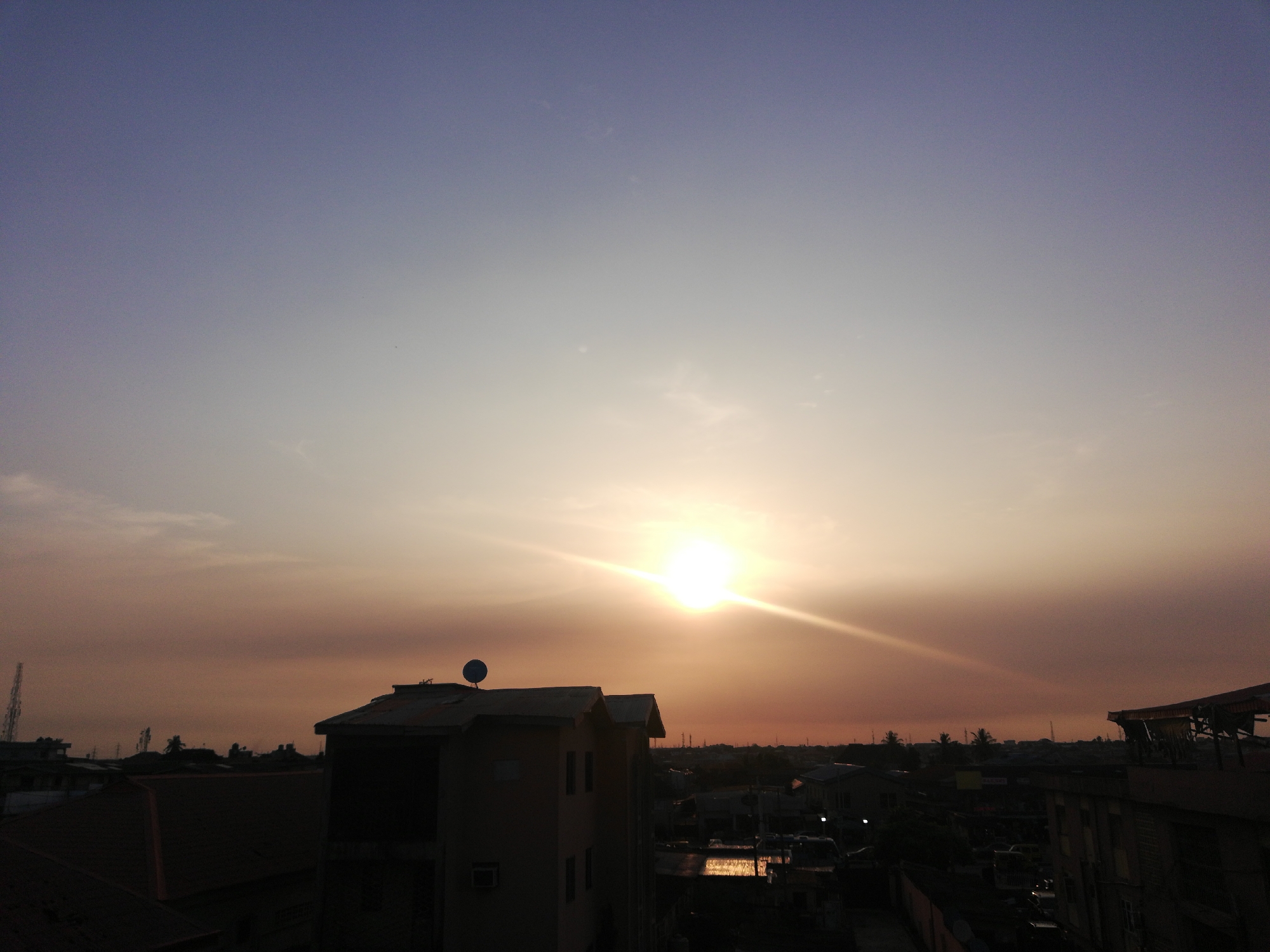 This image captures a vibrant sunset over an urban or suburban landscape, likely in Lagos, Nigeria, given the provided context. The sun is a dominant feature, positioned slightly to the right of the center, low on the horizon, and emitting powerful, bright rays that create a lens flare effect across the sky.
The sky transitions dramatically in color, from a deep purplish-blue at the very top, fading into lighter shades of blue and grey, before intensifying into warm golden-orange and light yellow hues around the sun and towards the horizon. Faint, wispy clouds are visible in the upper parts of the sky, adding subtle texture. The overall weather appears clear, with possibly some atmospheric haze enhancing the warm tones of the sunset.
The lower half of the image is dominated by the silhouetted rooftops and buildings of a dense urban environment, darkened by the backlighting from the setting sun. In the mid-ground, a prominent two-story building features a dark roof and a satellite dish, with hints of its facade and windows visible. Other structures, including what appears to be a communication tower or mast on the far left, are largely indistinct dark forms against the colorful sky. Scattered palm-like trees are also discernible as dark outlines among the buildings. No specific human activity is visible, and the scene appears calm at the end of the day. There is no visible text in the image. The composition strongly emphasizes the beauty of the sunset contrasting with the shadowed cityscape below, typical of metropolitan areas at dusk.