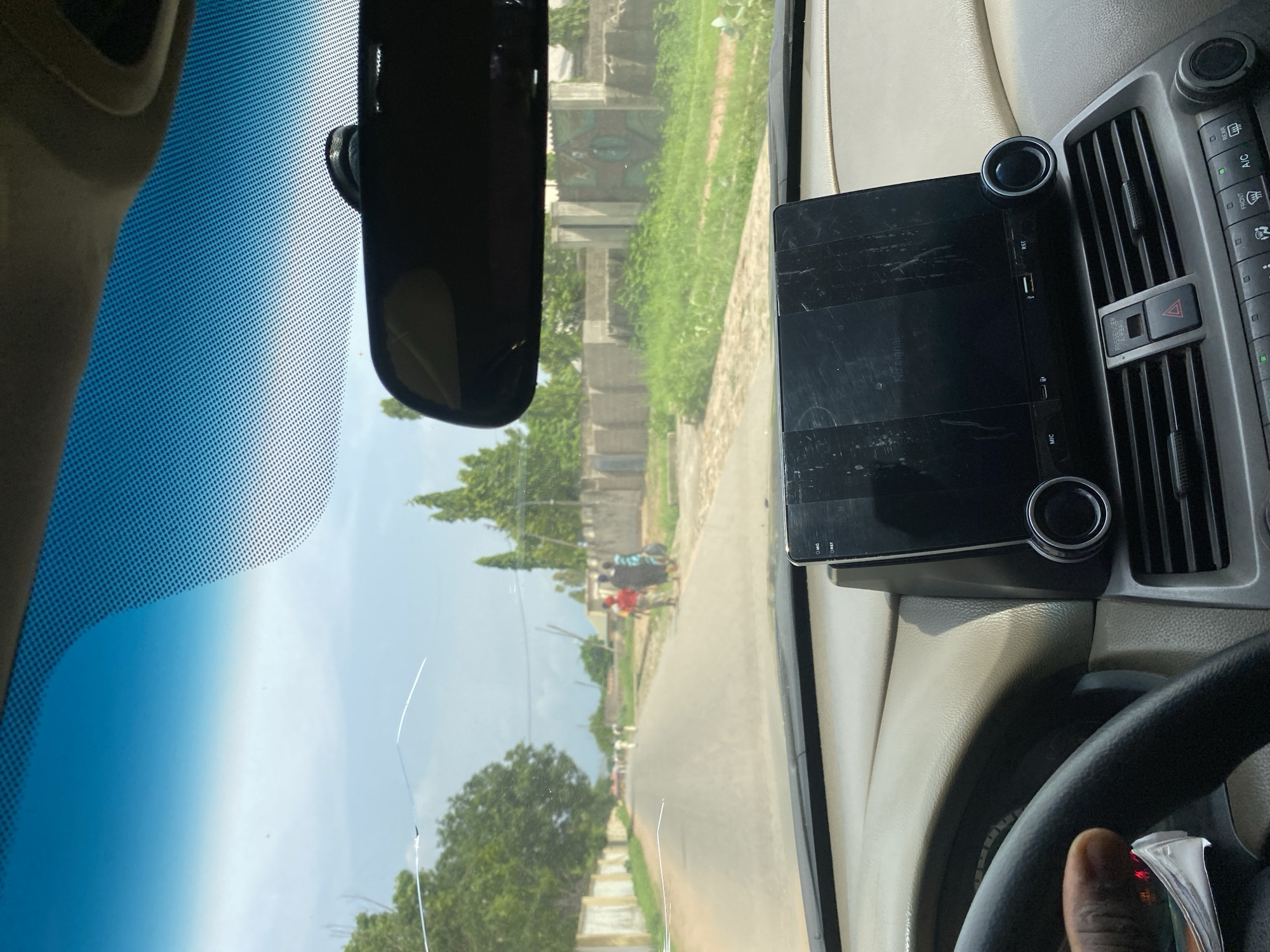 The image is taken from inside a car, looking out through the windshield. The windshield has visible cracks. Outside the car, a paved road stretches into the distance, lined on both sides by walls and vegetation. Several individuals, appearing to be adults, are walking along the road. The scene suggests a daytime setting with natural lighting. The context of Howayi, Nigeria, indicates an urban or semi-urban environment. The interior of the car shows a dashboard with a central display screen, part of a steering wheel, and air conditioning vents.