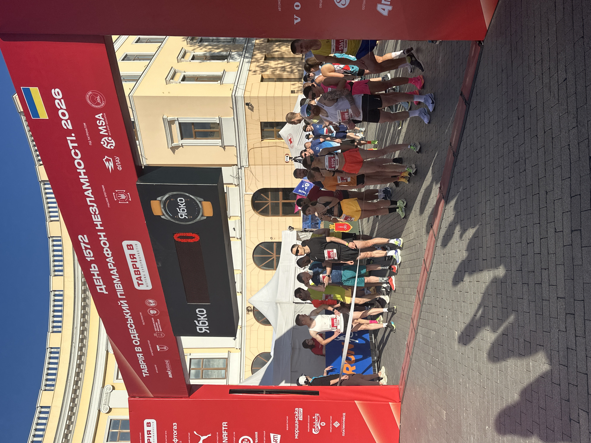 The image shows the start/finish line of a marathon in Odesa, Ukraine. A large red banner stretches across the frame, displaying event information in Ukrainian, including "ДЕНЬ 1572" (Day 1572) and "НЕЗЛАМНОСТІ. 2026" (Indomitability. 2026), along with sponsor logos. Below the banner, a digital display shows the word "Ябко" (Apple) and a timer displaying "0". A group of runners, both male and female, are lined up at the start, some wearing race bibs with numbers such as 5579, 5570, 5375, 5089, and 5361. They are dressed in athletic wear, including shorts and tank tops. Some runners are looking at their phones or stretching. A white tent is visible in the background. The ground is paved with cobblestones, and shadows of the runners are cast across it. The architecture in the background suggests a historic building with yellow walls and arched windows, characteristic of Odesa. The sky is clear and blue.
