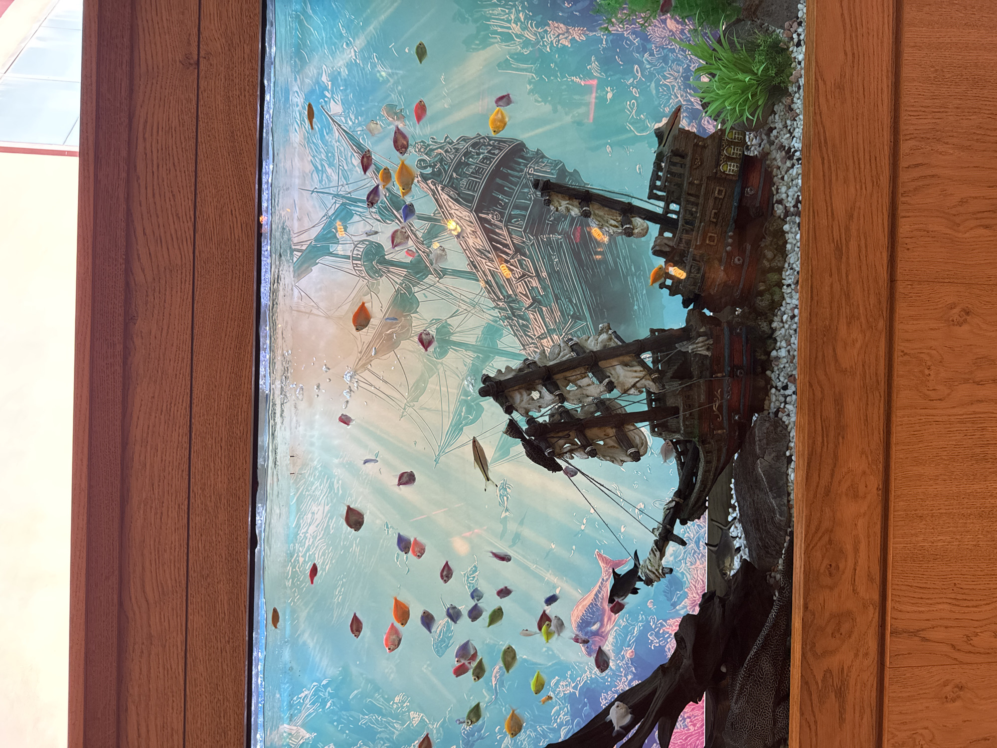 The image shows a large, built-in aquarium with a wooden frame. Inside the aquarium, there's a decorative shipwreck model and various artificial plants and gravel. Many small, colorful fish swim within the aquarium. The background of the aquarium features an underwater scene, potentially a printed backdrop. The aquarium is framed by light brown wood on both sides. The precise location within Plovdiv, Bulgaria, cannot be determined from the visual information provided.