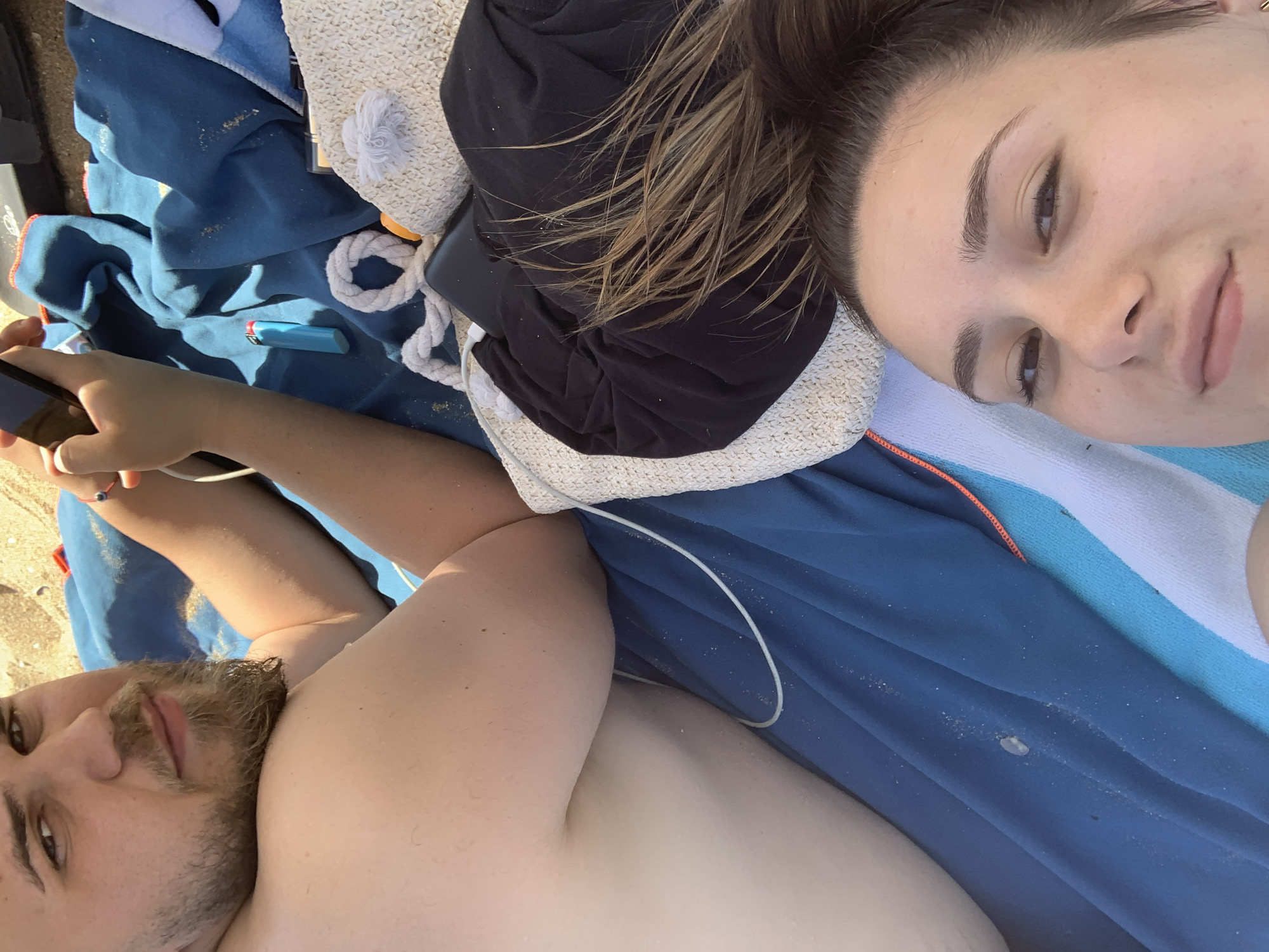 This is a close-up, high-angle, outdoor, selfie-style photograph taken on a beach in Sozopol, Bulgaria. The scene is dominated by a blue towel, sand, and various personal items. In the foreground, the face of a young woman is visible, her hair is pulled up, and she is looking towards the camera. To the left and slightly behind her, a man's face and shoulder are in view. He has a beard and is looking away from the camera, holding a smartphone. The man is partially covered by a blue towel.
Scattered on the towel are several objects: a blue lighter, a white woven bag with a rope handle and a white pom-pom, a black garment, and what appears to be a charging cable. The sand is visible around the towel. The lighting suggests it is daytime, likely sunny. The overall impression is a relaxed, casual scene of two people enjoying time on the beach.