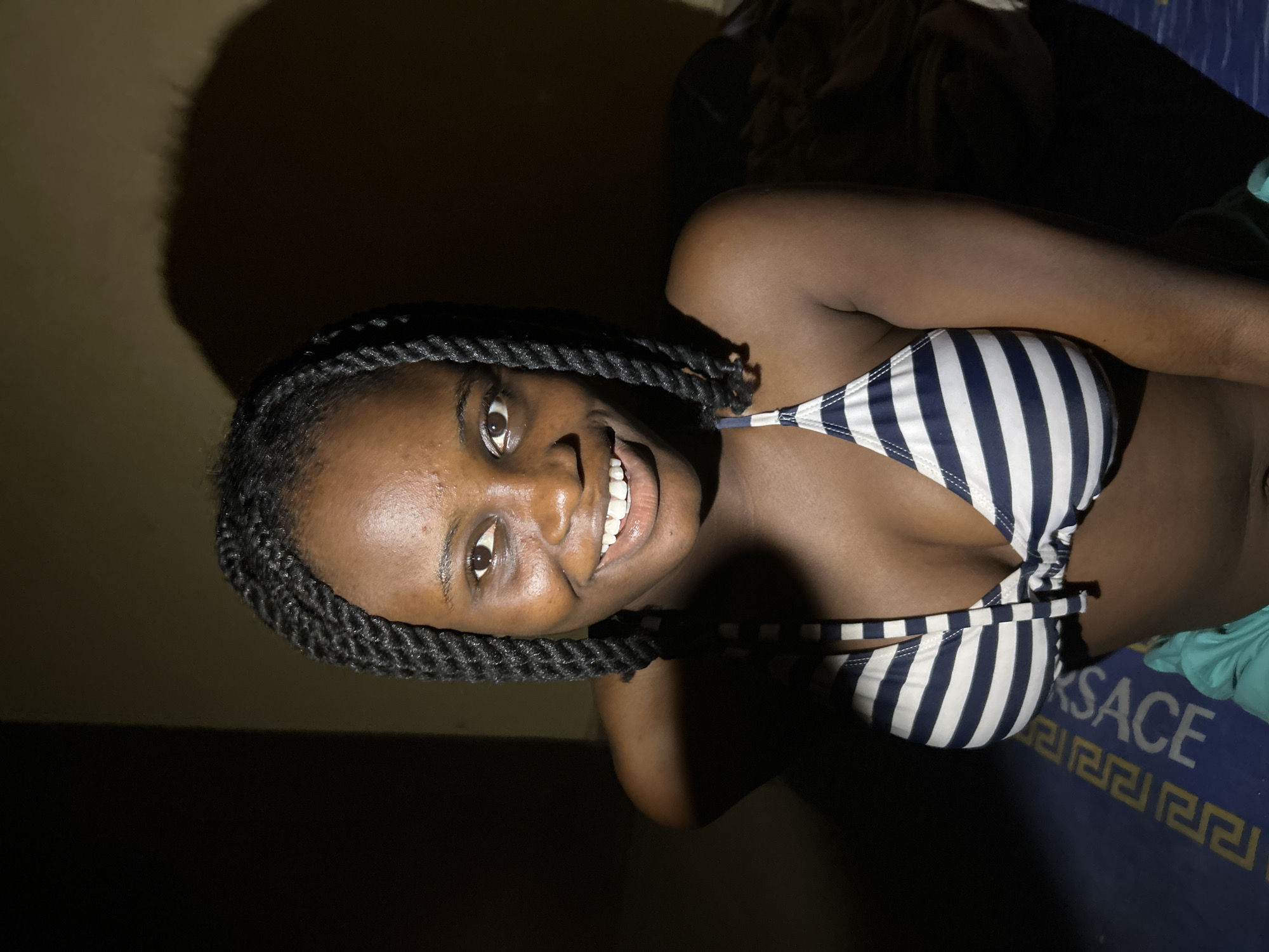 In Ihiagwa, Nigeria, an individual is captured in a medium shot, looking up and to the right. The subject has dark skin, braided hair styled into two thick plaits on either side of their head, and is wearing a blue and white striped bikini top. Their facial expression is a wide smile, revealing their teeth. The background is dark and appears to be an indoor setting, with a shadow cast on the wall behind the subject, suggesting a single light source. A portion of a blue mat with a yellow Greek key pattern and text is visible at the bottom of the frame.