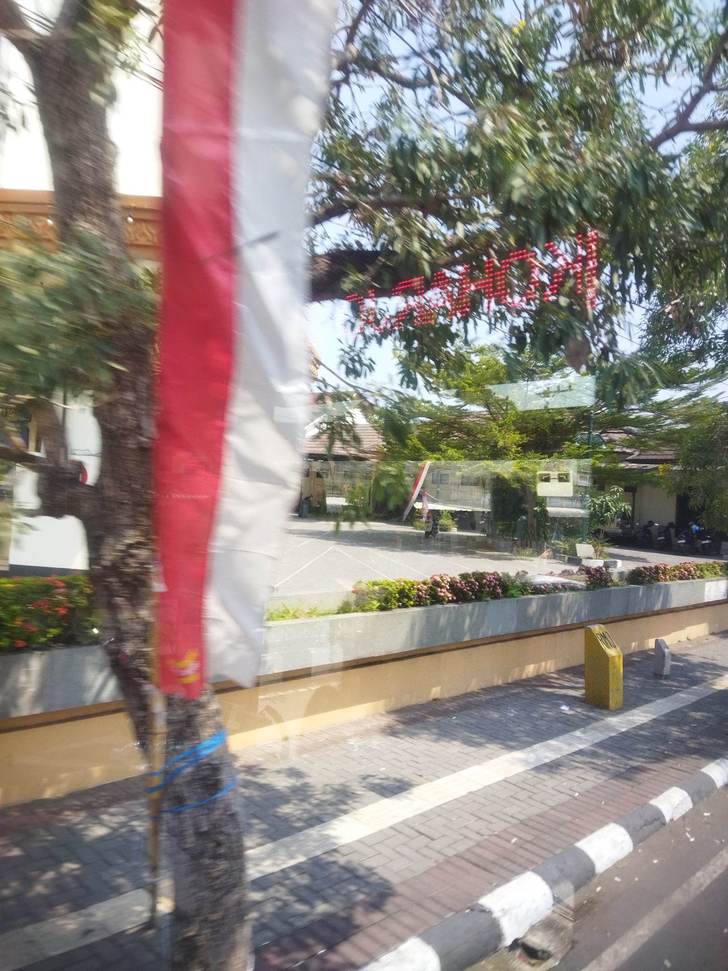 This is a slightly blurry, eye-level, outdoor shot taken from a moving vehicle, likely a bus or car, in Sukoharjo, Indonesia. The image captures a street scene with several key elements.
In the foreground, a prominent, vertical Indonesian flag, consisting of red and white stripes, is positioned to the left of the frame. The flag appears to be attached to a tree trunk, which is partially visible and bound with blue rope. The tree is growing on the left side of the image, with its branches and leaves extending upwards and across the frame.
To the right of the flag, a sidewalk is visible, paved with rectangular bricks in a herringbone pattern. The sidewalk is bordered by a curb painted with black and white stripes. A yellow marker post is placed on the sidewalk.
Behind the sidewalk and a low wall, a paved area is visible, possibly a parking lot or courtyard. In this area, there are several buildings with tiled roofs, appearing to be shops or residences. Some of the buildings have a modern architectural style. There are also trees and greenery around the buildings. A few motorcycles are parked in the background.
Above the buildings, a bright blue sky is visible, with some scattered clouds. A red LED sign, with an unreadable message, is illuminated and partially obscured by the tree branches. The overall impression is that of a sunny day in an urban or semi-urban environment. The blurring suggests movement, indicating the photo was taken while in transit.