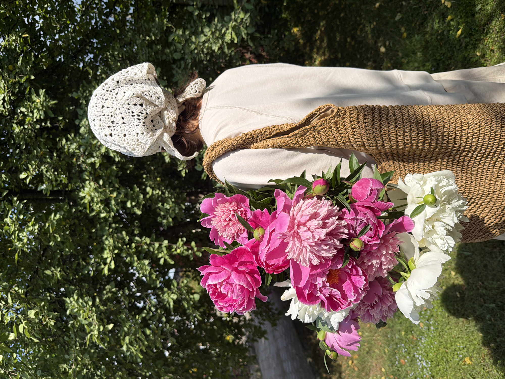 A person seen from behind, wearing a light-colored sweatshirt and a white crocheted headscarf, holds a large bouquet of pink and white peonies. The person is also carrying a woven straw bag over their right shoulder. They are standing outdoors, with a backdrop of lush green trees and foliage. The ground is covered in green grass with scattered fallen leaves. The lighting suggests it is a sunny day, casting shadows on the grass. The scene appears to be set in a garden or park, possibly in the Прикамский region of Russia given the context.