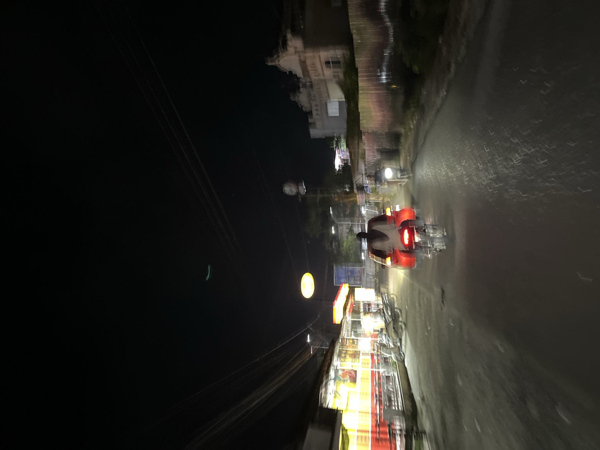 This is a nighttime, eye-level, wide-angle shot taken from a moving vehicle on a paved road in Langsa, Indonesia. The scene depicts a street at night with several elements indicating a commercial or residential area.
On the left side of the road, there is a building with bright yellow and red lighting, likely a shop or restaurant, with tables and chairs visible outside. Several motorcycles are parked in front of this establishment. Overhead power lines crisscross the sky.
In the center of the road, a person is riding a motorcycle with a red taillight and illuminated by the motorcycle's headlight. Another motorcycle is visible further ahead on the right side of the road, also lit by its headlight.
On the right side of the road, there is a long, corrugated metal fence. Behind the fence, a building with architectural details, possibly historical, is partially visible. Further down the road, more lights suggest other buildings or businesses.
The road surface appears wet, reflecting the lights from the surrounding environment. The overall impression is a bustling street scene at night, with activity and illumination despite the darkness. The image captures a sense of movement, given the blurriness of some objects, suggesting it was taken while the camera was in motion.