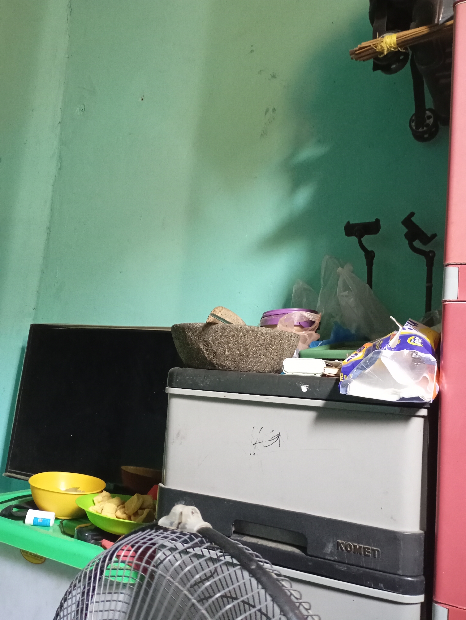 In a room with a light green wall, a large black television is placed to the left. On top of a gray plastic cabinet, a stone mortar and pestle are visible. The cabinet has "KOMET" embossed on its lower front. To the right of the cabinet is a tall pink storage unit. In the foreground, a partially visible fan with a wire grill is positioned on the floor. Various items, including bowls of food, a small plastic container, and plastic bags, are scattered on the green surface to the left of the cabinet. The overall scene suggests a domestic setting in Kecamatan Haurgeulis, Indonesia.