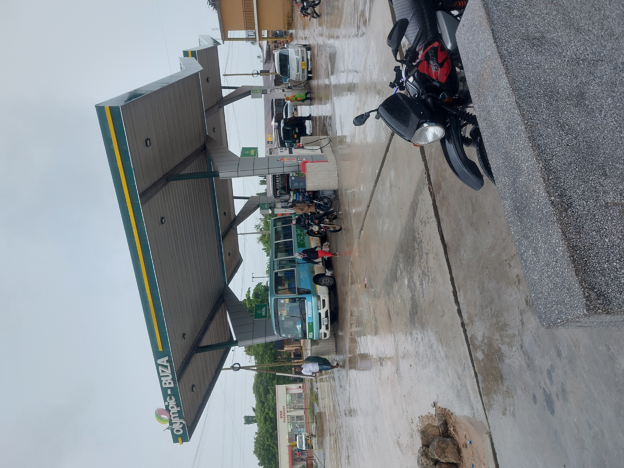 The image depicts a gas station in Dar es-Salaam, Tanzania, named "Olympic-Buza." There is a light-blue minibus being refueled, with people standing near the vehicle. Several motorcycles are parked near the gas pumps, and other vehicles, including a white van and a black car, are also at the station. The ground is wet, suggesting it has recently rained. A man is seen holding an umbrella. The surrounding area includes a pharmacy ("BRK Pharmacy") and other buildings.