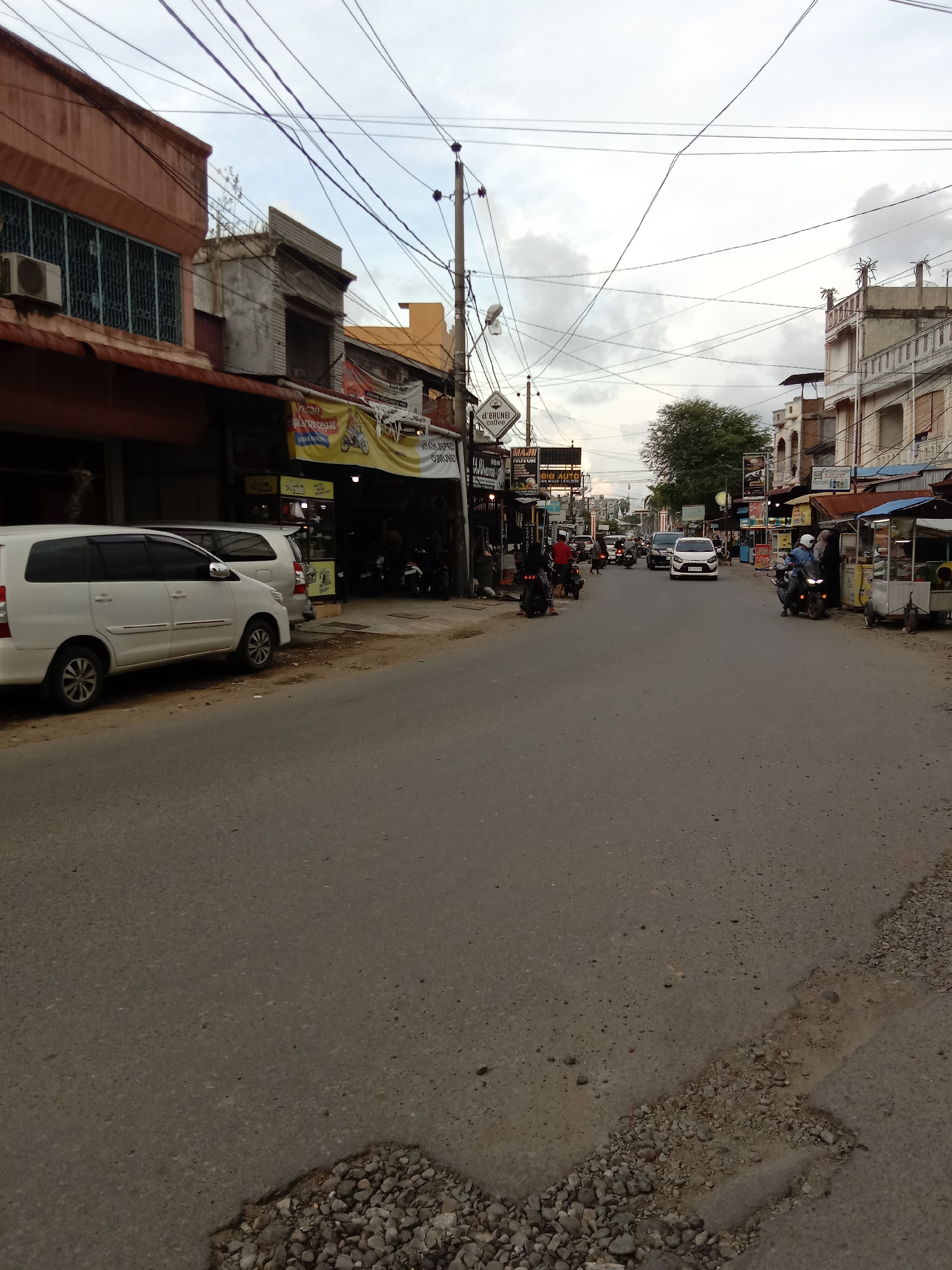 The image shows a street scene in Lhokseumawe, Indonesia. The street is paved, with visible wear and tear, including a patch of gravel in the foreground. Several buildings line both sides of the street. The buildings are a mix of architectural styles, some with awnings or verandas. Power lines crisscross above the street.
Vehicles are present, including cars and motorcycles. Some motorcycles are parked along the side of the road, while others are in motion. People are visible riding the motorcycles. There is a food stand on the right side of the street, and banners or signs hang from some of the buildings. The sky is overcast.