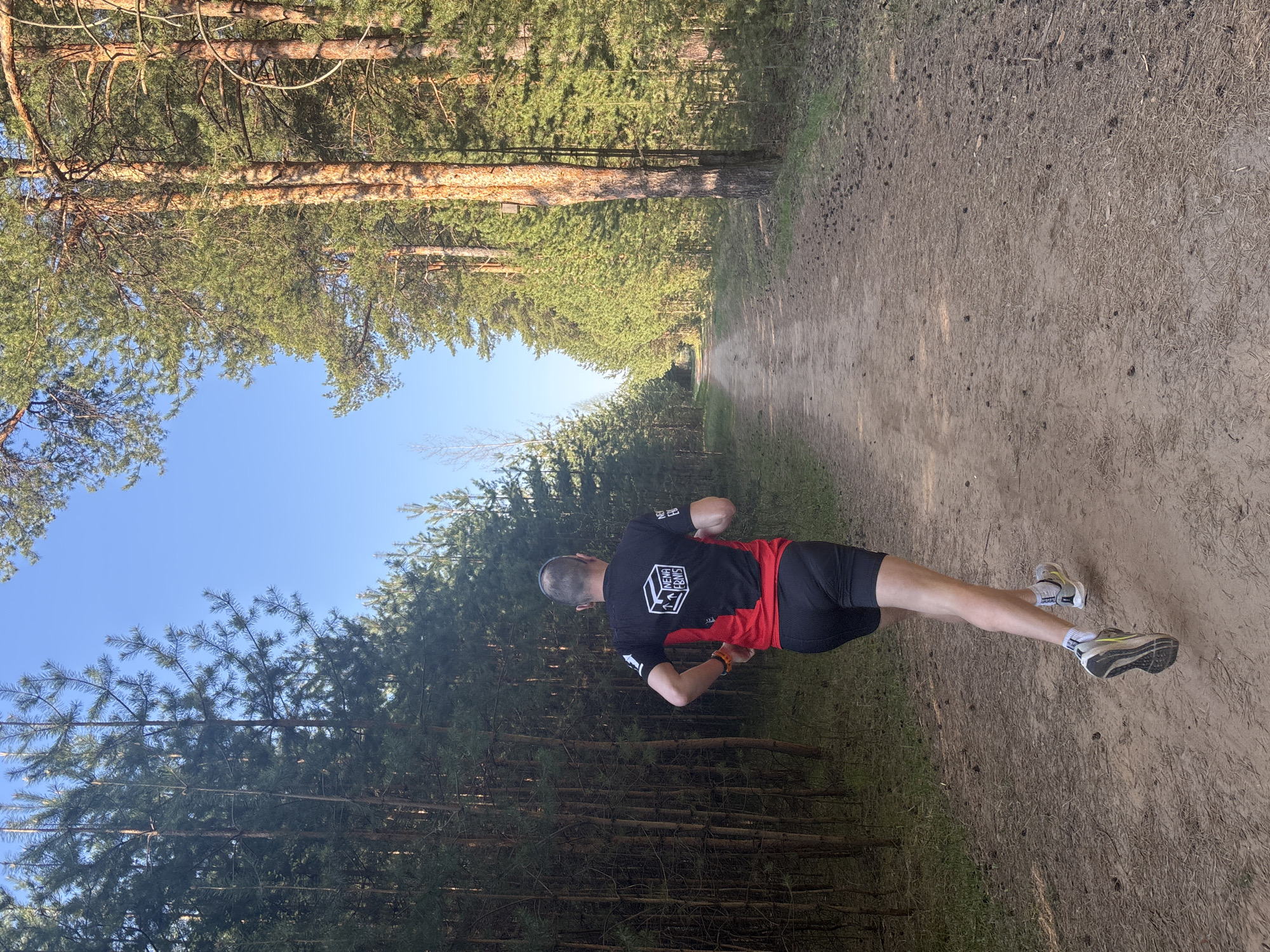 The image shows a man running on a dirt path through a forest. The trees are tall and green, and the sky is blue. The runner is wearing a black and red shirt, black shorts, and running shoes. The location is likely a forested area near the city of Chaikovsky, Russia, given the presence of coniferous trees and the description provided. The man appears to be in motion, his arms bent and legs extended in a running stride. The environment seems natural with a path.