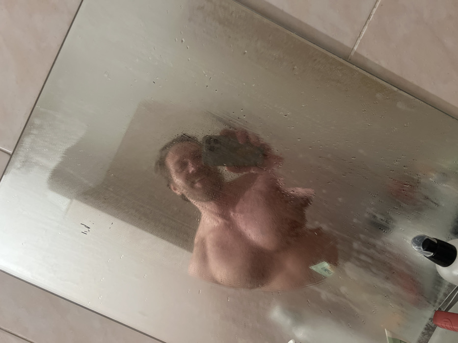 A man with light brown hair is reflected in a foggy mirror, holding a cell phone. He appears shirtless. Water droplets are visible on the mirror's surface. The mirror's frame is light-colored, suggesting a bathroom setting. Various toiletries are situated at the bottom right corner of the mirror. The overall tone of the image is intimate and informal, capturing a self-portrait style.