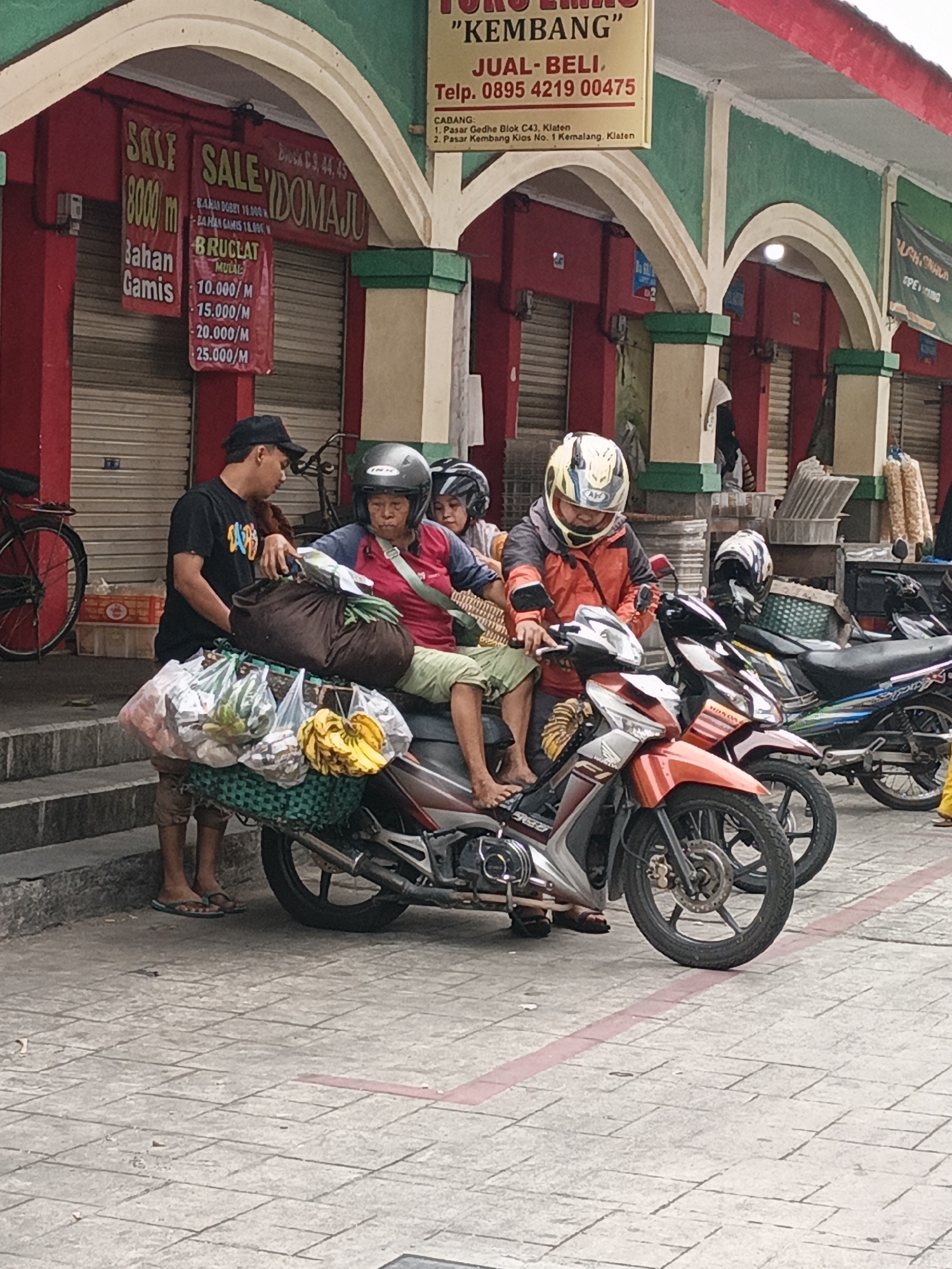 In Klaten, Indonesia, three people are gathered around a motorcycle laden with goods. A man wearing a black t-shirt and a black cap stands on the left, facing the motorcycle and appearing to interact with another person. To his right, a woman wearing a red shirt and a helmet is seated on the motorcycle, holding a bag. Behind her, another person wearing a helmet is partially visible, also seated on the motorcycle. The motorcycle is overloaded with various bags and produce, including bananas. The scene takes place in front of shops with red and green facades, and a large banner advertising "SALE" and prices is visible on the left. The ground is paved with square tiles.