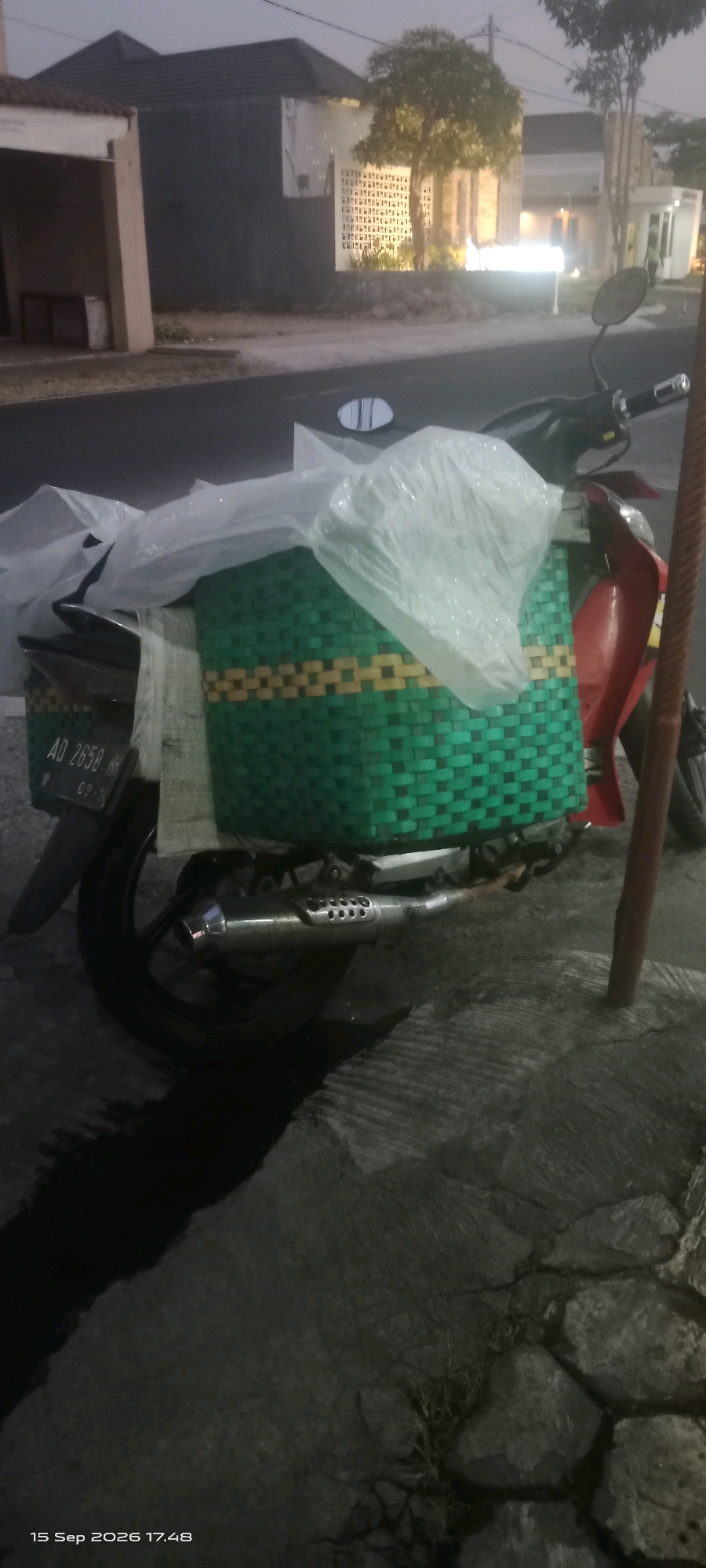 The image depicts a motorcycle parked on a stone-paved surface near a road. The motorcycle is equipped with a green and yellow woven basket attached to its rear, covered with a translucent plastic material. The license plate of the motorcycle displays "AD 2658 HH". In the background, buildings with illuminated windows and trees are visible, suggesting an urban or suburban environment. The lighting indicates it is either dawn or dusk. A red pole stands to the right of the motorcycle. The timestamp "15 Sep 2026 17:48" is present in the lower left corner, suggesting a potential recording date. The location is identified as Merbung, Indonesia.