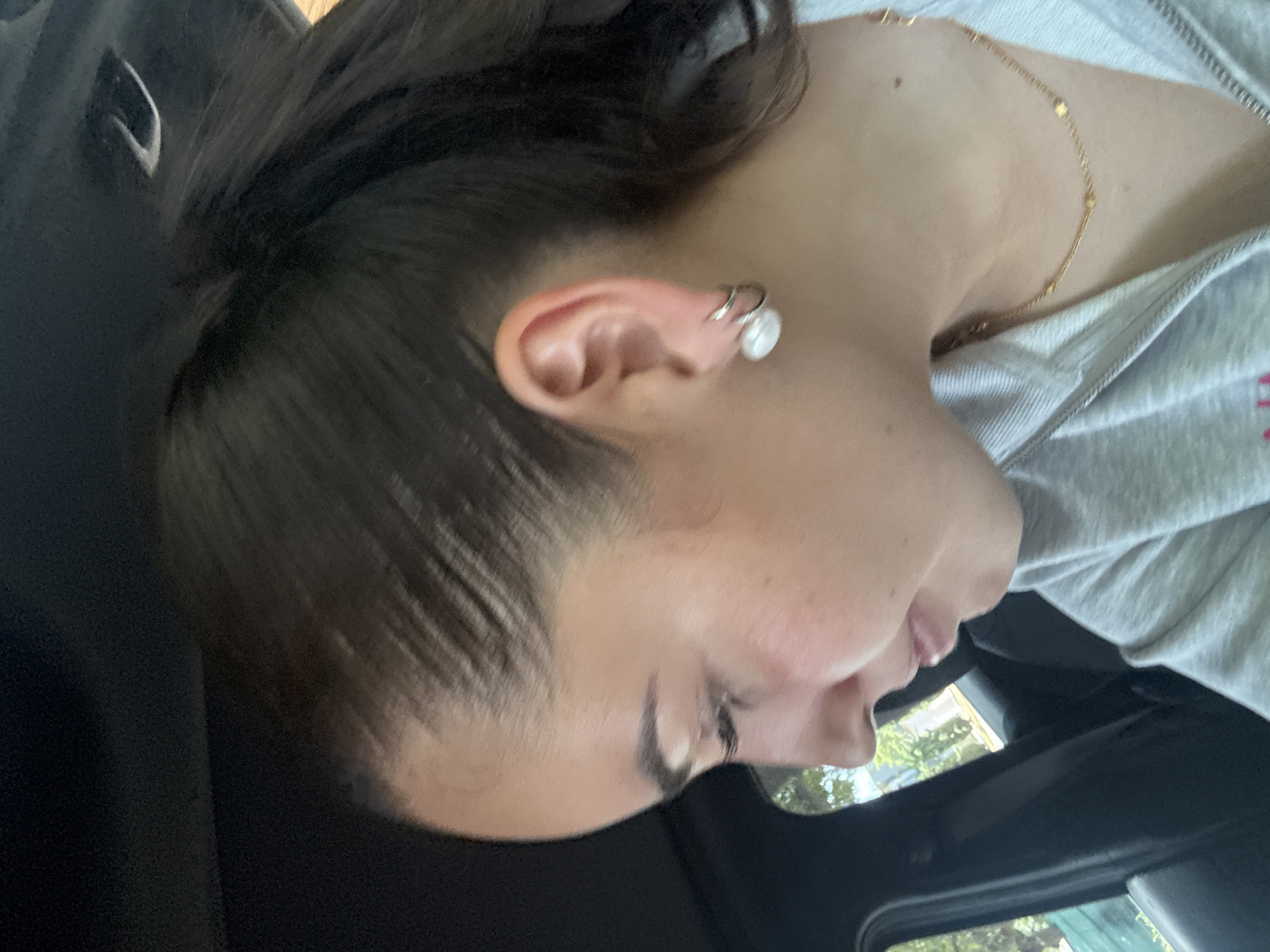 The image, rotated 90 degrees clockwise, presents a close-up profile of a person, likely a woman, from the right side of her head, neck, and upper chest. Her dark hair is pulled back, revealing her ear, which is adorned with a prominent pearl earring and a silver-toned ear cuff. A delicate gold-colored necklace with small beaded details is visible around her neck, resting on the fabric of a light gray zipped hoodie.
The person's face is angled downwards, with her eyes appearing closed or cast down, suggesting a moment of contemplation, rest, or looking at something below the frame. Her lips are slightly parted. The background on the left is a dark, indistinct car interior, possibly a headrest or seat. To the right, through a car window, blurred outdoor scenery is visible, including green foliage and what appears to be a building structure, indicating that the photo was taken during the daytime. The lighting suggests it is a bright or sunny day.
No discernible text is visible on any subject or object within the frame. The overall scene conveys a calm atmosphere, possibly from inside a vehicle in San Bernardino Tlaxcalancingo, Mexico.