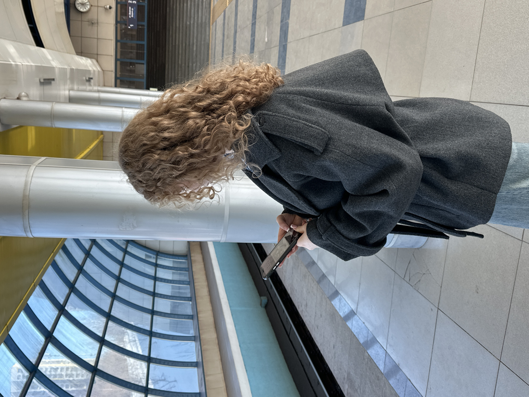 The scene depicts a metro station platform in the city of Vorob'evka, Russia, characterized by an arched glass roof structure. A female individual, seen from a slightly elevated rear-right perspective, occupies the foreground. She has long, curly, light brown hair and is wearing a dark grey, oversized coat with distinctive shoulder flaps and a tie waist. Her lower garment is light blue jeans. She stands facing towards the platform edge and tracks, holding a black smartphone horizontally with both hands, interacting with its screen which displays the time "12:54." A silver ring is visible on her right ring finger, and her fingernails are manicured with a light polish.

The platform floor is comprised of light grey tiles, some exhibiting visible cracks and wear. A dark grey safety strip runs along the platform edge. Below the platform, two subway tracks are visible on a dark substrate, separated by a light blue wall.

The station's architecture includes multiple cylindrical, light-colored support columns. Above these columns, a bright yellow structural beam is discernible, beneath a predominantly white/grey ceiling. The most prominent feature is the extensive, arched glass roof, framed by dark blue/grey supports, allowing natural light to illuminate the station. Through the glass, a snow-covered exterior environment with distant buildings is visible.

In the mid-ground, further along the platform, a set of dark stairs leads upwards. A large, circular analog clock with a white face and dark numerals is mounted on a far wall, near what appears to be a directional sign with Russian text and numbers (e.g., "П-2-3"). Overhead lighting fixtures are integrated into the station's ceiling design.
