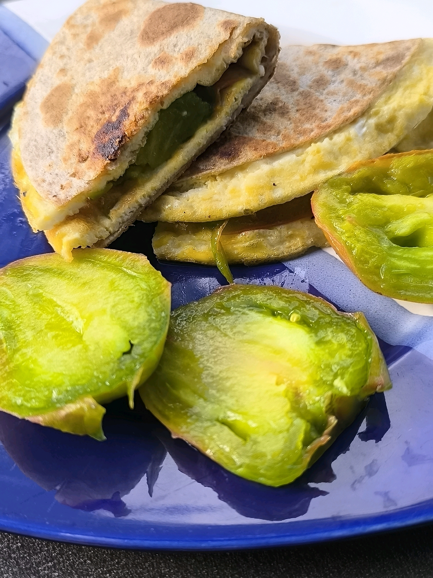 A close-up, overhead shot shows a half-eaten quesadilla with green filling, possibly avocado, and melted cheese, served on a blue and white patterned plate. Slices of green bell pepper are arranged around the quesadilla. The scene is brightly lit, with shadows cast on the plate. The background is blurred, suggesting an outdoor setting. The plate is positioned on a dark, textured surface, possibly a table or placemat. No specific landmarks or city references can be identified. The overall impression is a casual meal or snack.