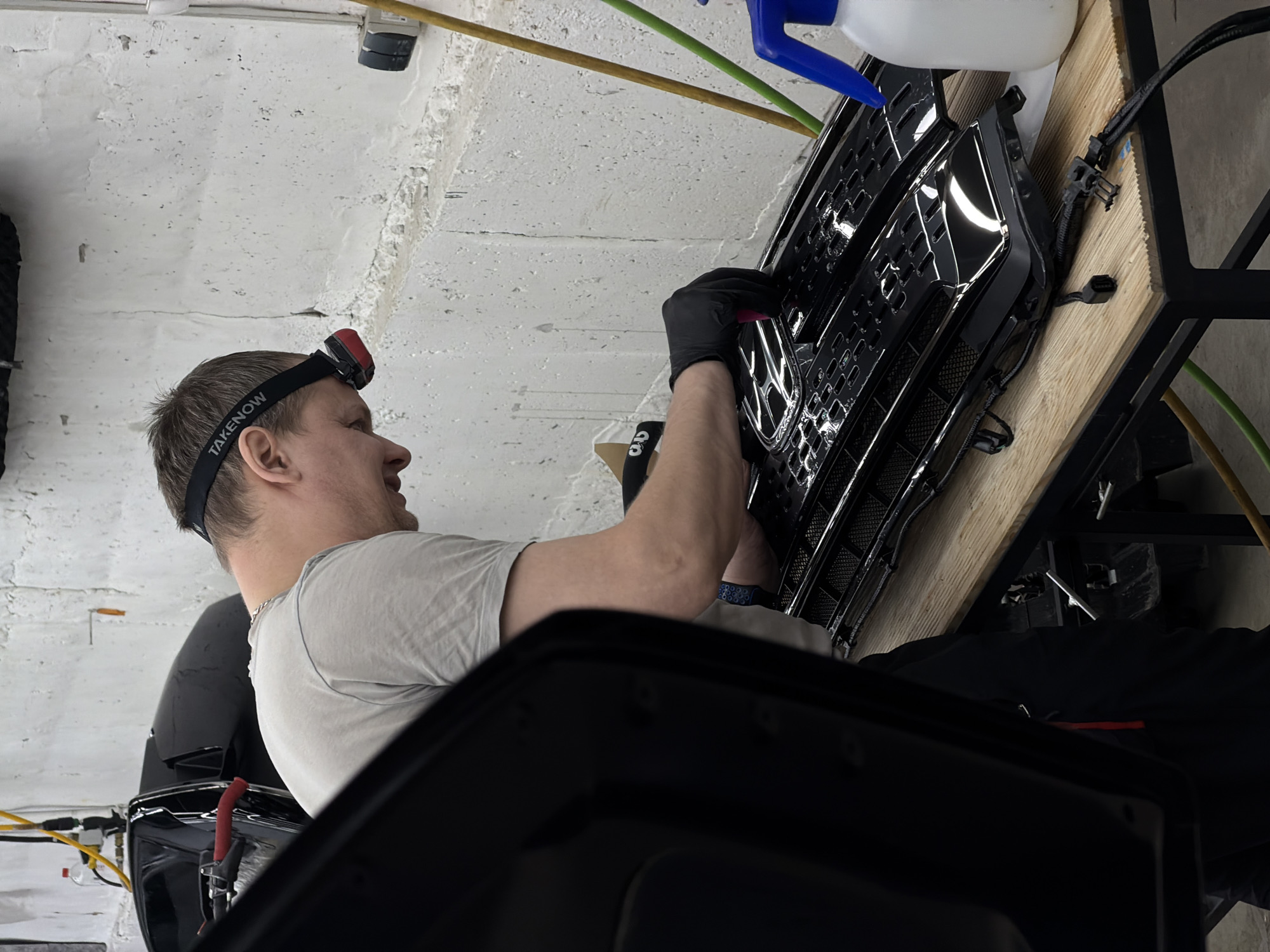 An image, captured with a rotated perspective, depicts a man diligently working on a car grille in what appears to be a workshop or garage in Krasnoyarsk, Russia. The man, seen from the side, wears a light-colored short-sleeved t-shirt and black gloves on his hands. A headlamp with the brand name "TAKENOW" visible on its strap is worn on his head, suggesting detailed work or low light conditions.
He is focused on a glossy black car grille, which features a prominent honeycomb-like pattern. His gloved hands are manipulating a small red tool or object within the intricate design of the grille, possibly during an installation, repair, or detailing process. The grille rests on a light-colored wooden surface, likely a workbench or table, with black wiring harnesses connected to it.
In the background, a white spray bottle with a blue nozzle and various industrial-style hoses (green and yellow) are visible. The walls and ceiling are a rough, textured white or light gray, resembling concrete. A light fixture or security camera is mounted overhead. The overall setting indicates an indoor, practical workspace. The man appears concentrated on his task.