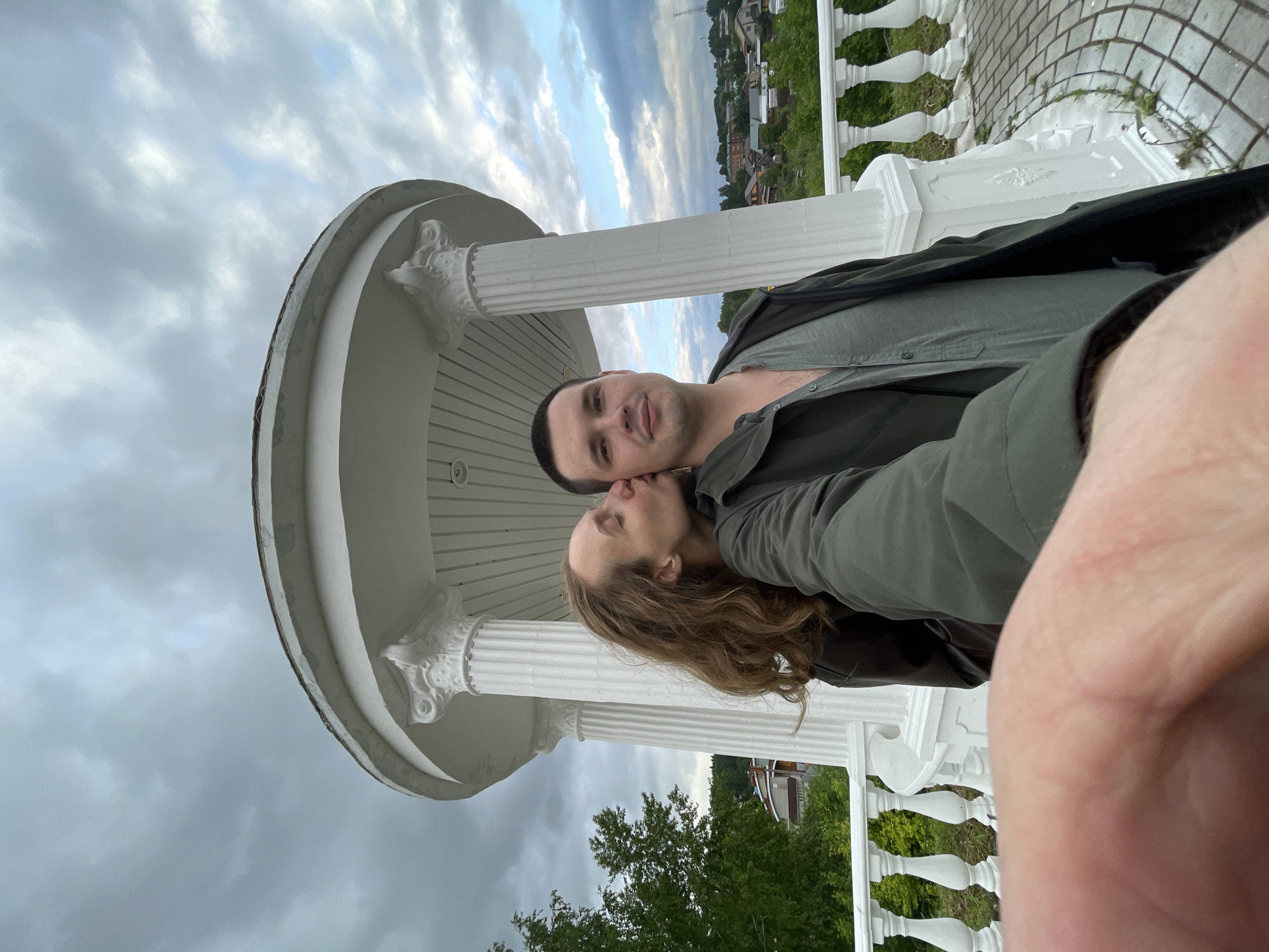 A selfie captures two individuals in a white, classical-style rotunda or gazebo featuring fluted columns with Corinthian capitals and a rounded roof. The person on the left, with long brown hair, is wearing a dark jacket and is depicted kissing the right cheek of the person on the right. This second individual, with short dark hair, wears a grey shirt under a dark green jacket and looks directly at the camera with a neutral expression. The bottom foreground shows a partial view of a hand holding the camera.
The rotunda is positioned on a paved surface with a circular pattern of grey stones, enclosed by a white balustrade. Beyond the balustrade, green deciduous trees are visible on the left. The background reveals an expansive view of a settlement comprising numerous houses and buildings, characteristic of a rural or village landscape. A tall communication tower is discernible in the far right background. The sky is largely overcast with grey clouds, though patches of lighter blue sky and white clouds are visible towards the right. The scene is located in Sharkan, Russia.