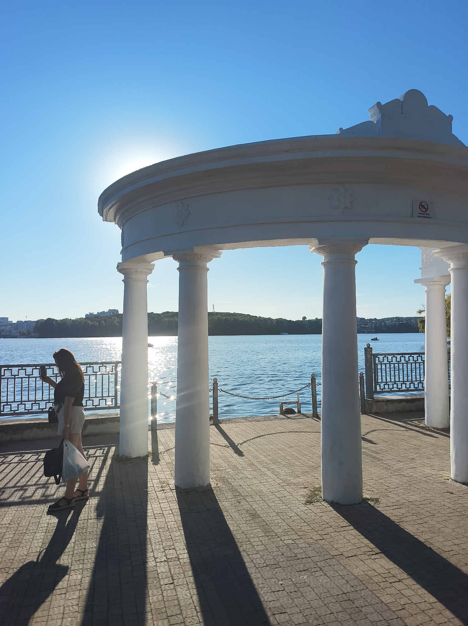 The image captures a sunny day by the Ternopil Pond in Ternopil, Ukraine. A woman stands to the left, holding a phone and a shopping bag, casting a long shadow. She is positioned on a paved walkway beneath a white, classical-style gazebo with fluted columns. The gazebo overlooks the water, with a railing separating the walkway from the pond. The water is a bright blue, reflecting the sunlight, and a few boats are visible in the distance. The background shows a tree-lined shore with buildings. The sky is a clear, vibrant blue.