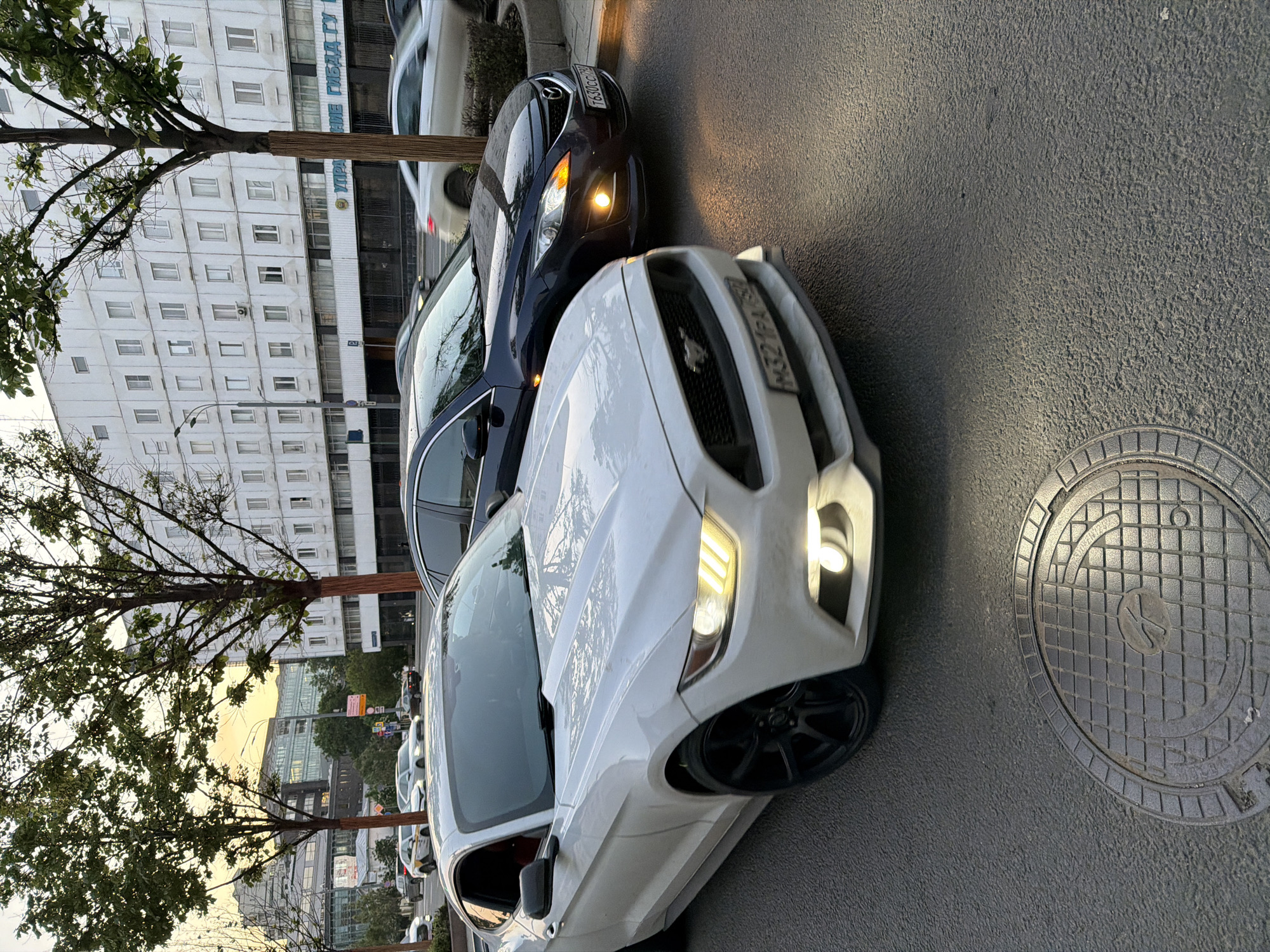The image shows two cars parked on the side of a street. The car in the foreground is a white Ford Mustang with its headlights on. Next to the Mustang is a dark-colored Mazda. Both cars have Russian license plates. To the left of the cars, there are buildings, trees, and street signs. In the background, there are more buildings and trees. The buildings appear to be in a city. A manhole cover is visible on the right side of the image. Based on the license plates and architectural style, the location is likely Moscow, Russia.